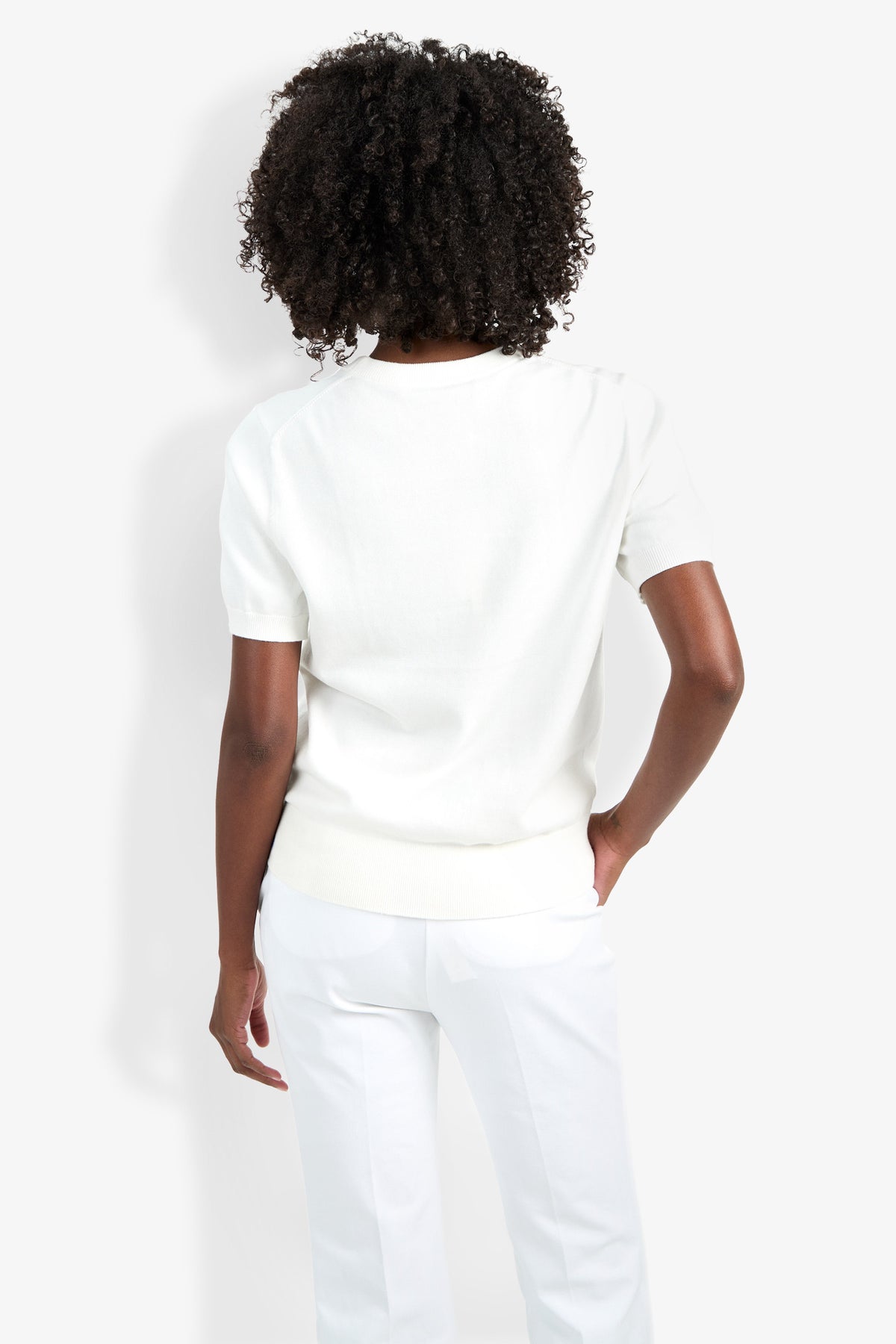 A person with curly hair is shown from behind, wearing the Maui Old Fashioned Sweater and white pants, standing against a plain light background with one hand in their pocket.