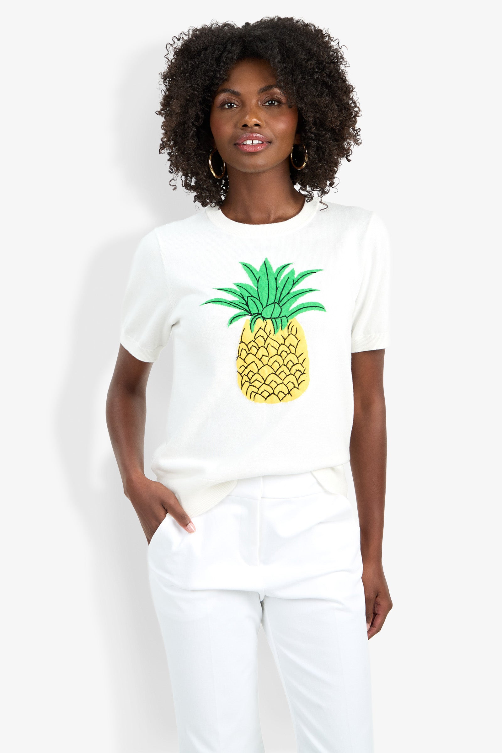 A woman with curly hair smiles slightly while wearing the Maui Old Fashioned Sweater— a white short-sleeve sweater featuring a bold yellow and green pineapple graphic—along with white pants, standing against a white background.