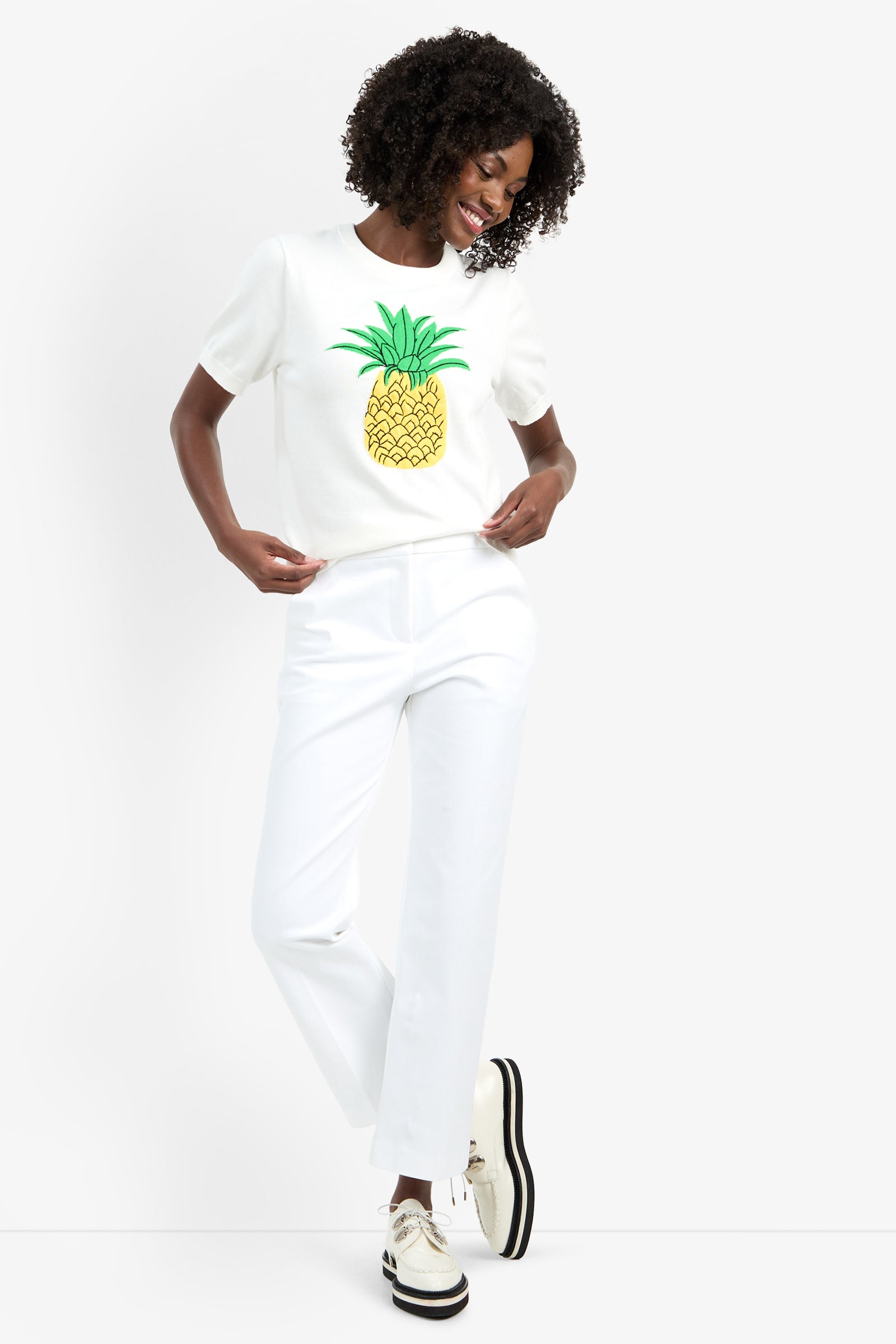 A person stands smiling against a plain white background, wearing the MAUI OLD FASHIONED SWEATER, white pants, and white platform shoes with black soles.
