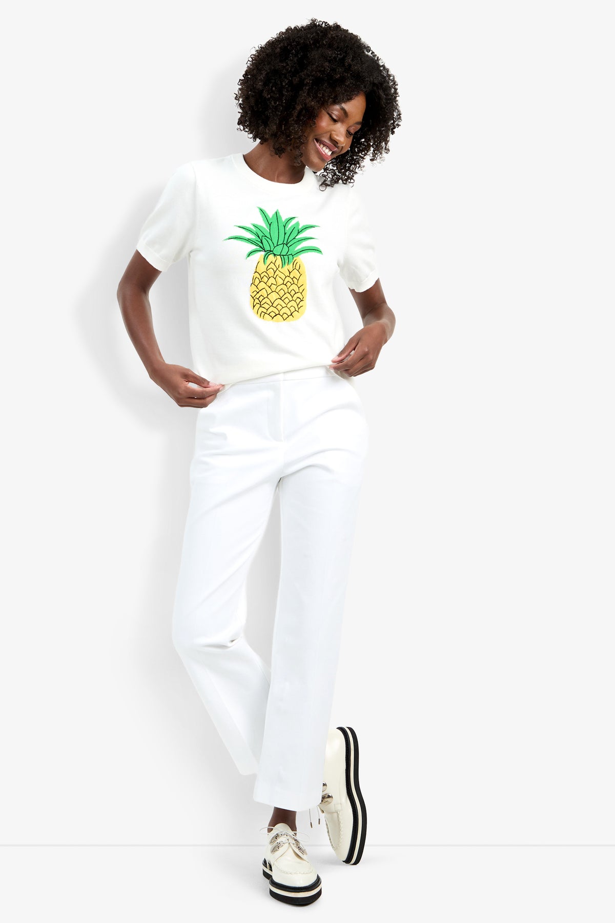 A woman smiles while wearing the Maui Old Fashioned Sweater, white pants, and cream platform shoes with black soles against a plain white background.