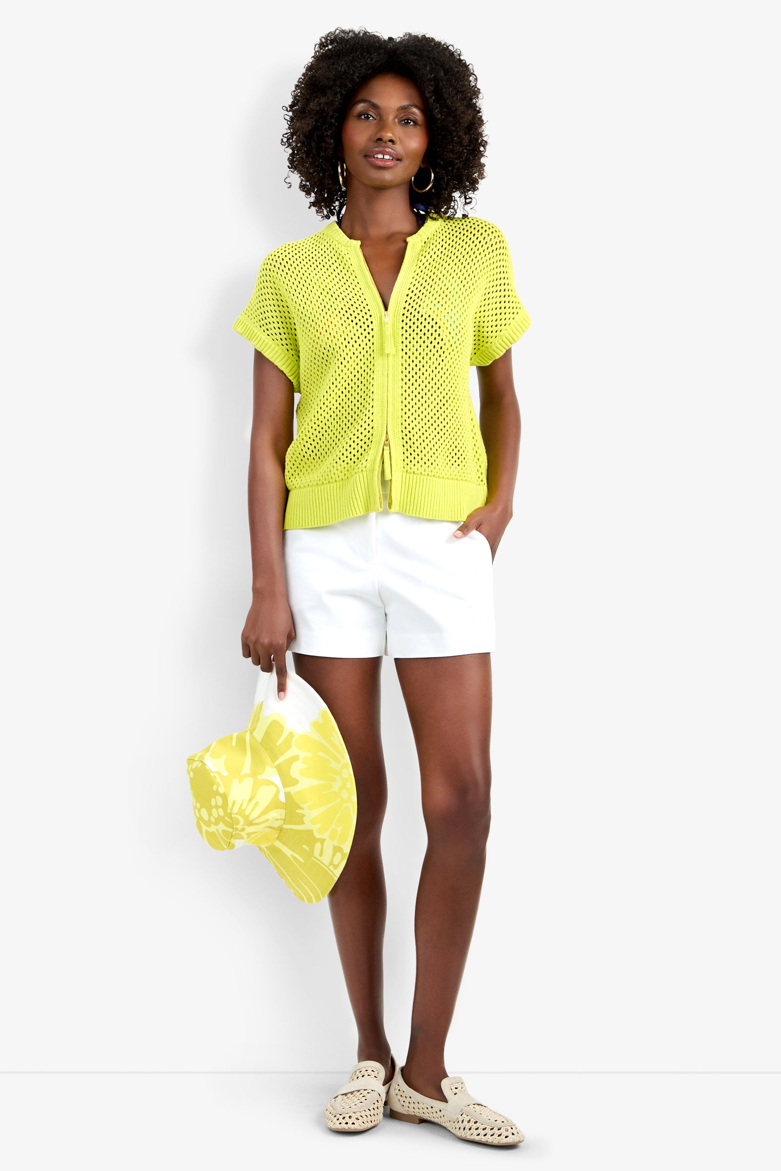 A woman stands against a white background, wearing the light yellow Hana Zip Cardigan over a yellow top, paired with white shorts, cream woven slip-on shoes, and holding a yellow floral bag.