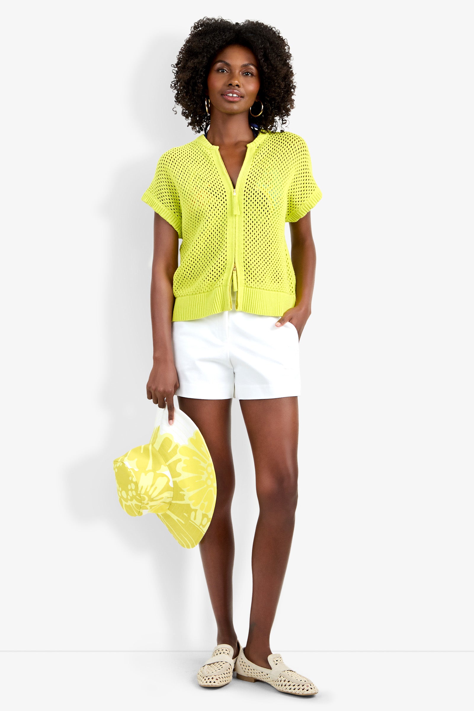 A woman with curly hair and blue earrings wears the yellow Hana Zip Cardigan over a yellow top, paired with white shorts, cream slip-on shoes, and carries a yellow floral tote bag against a white background.