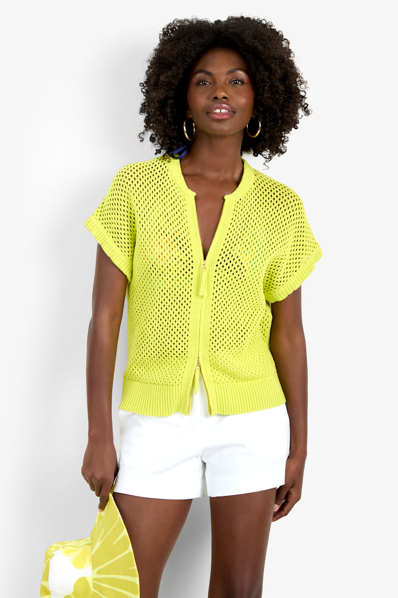 A woman with curly hair wears the HANA ZIP CARDIGAN, a bright yellow, short-sleeve, 100% cotton open-knit piece, over a top and white shorts. She holds a yellow and white patterned bag against a plain white background.