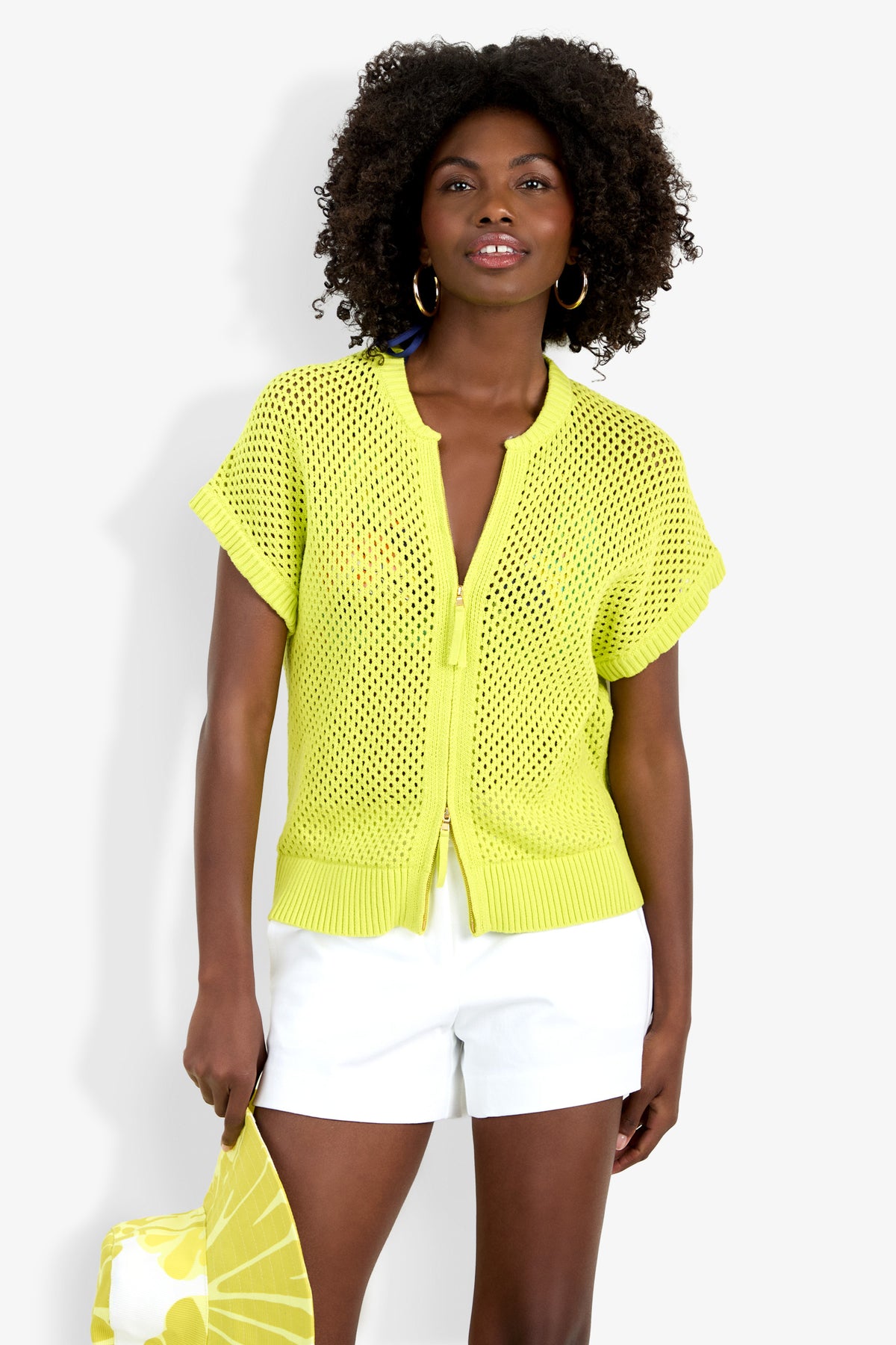 A woman with curly hair wears the bright yellow Hana Zip Cardigan over a blue top and white shorts, holding a matching yellow and white bag, standing against a white background.