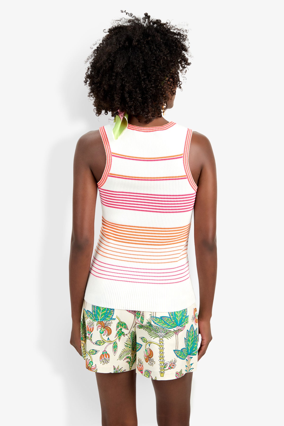 A person with curly hair, seen from behind, wears the Paolini Tank—a sleeveless white knit top with orange, pink, and red stripes—paired with light shorts featuring a colorful floral and leaf pattern.