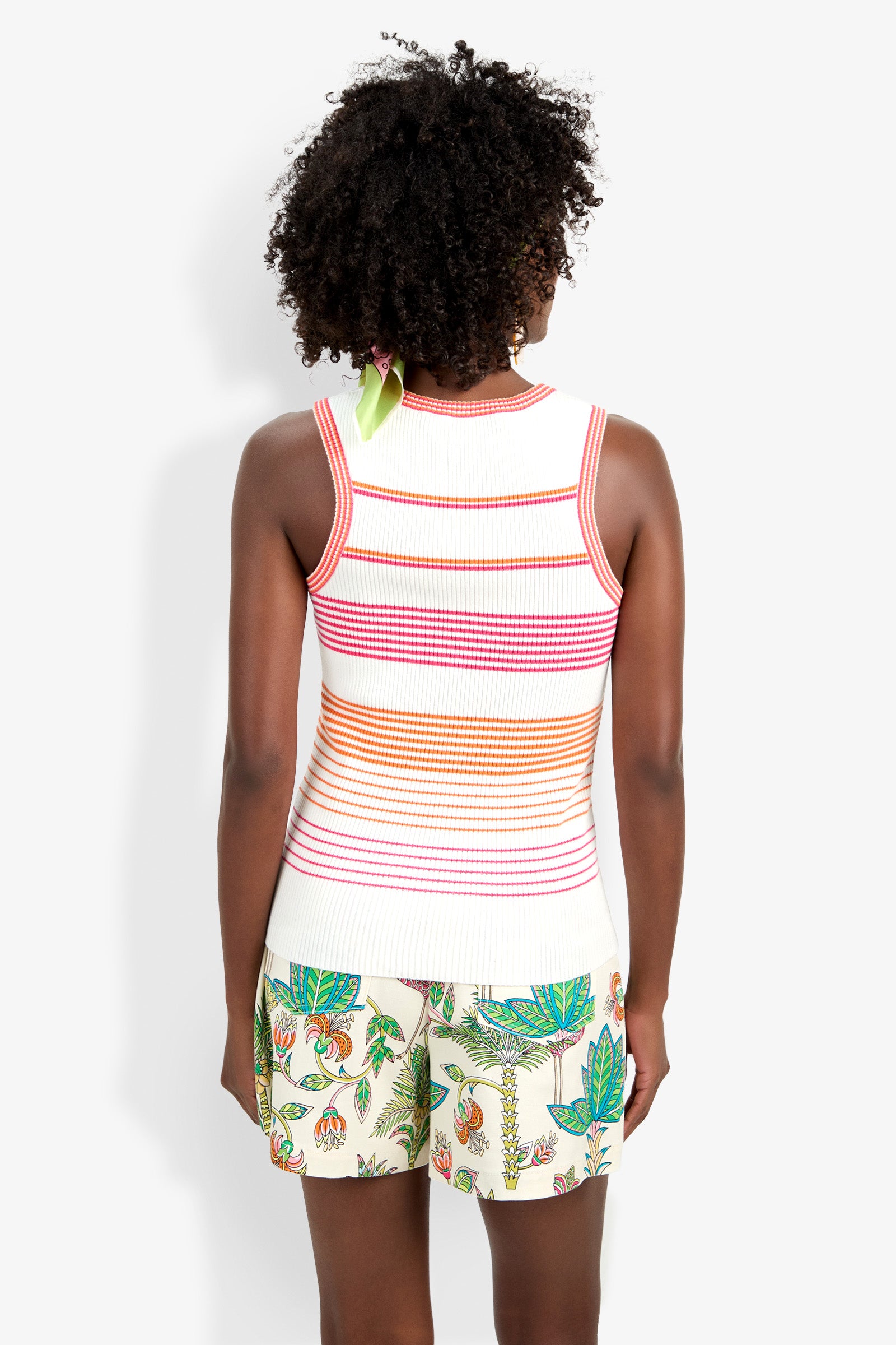 A person with curly hair, seen from behind, wears the Paolini Tank—a sleeveless white knit top with orange, pink, and red stripes—paired with light shorts featuring a colorful floral and leaf pattern.