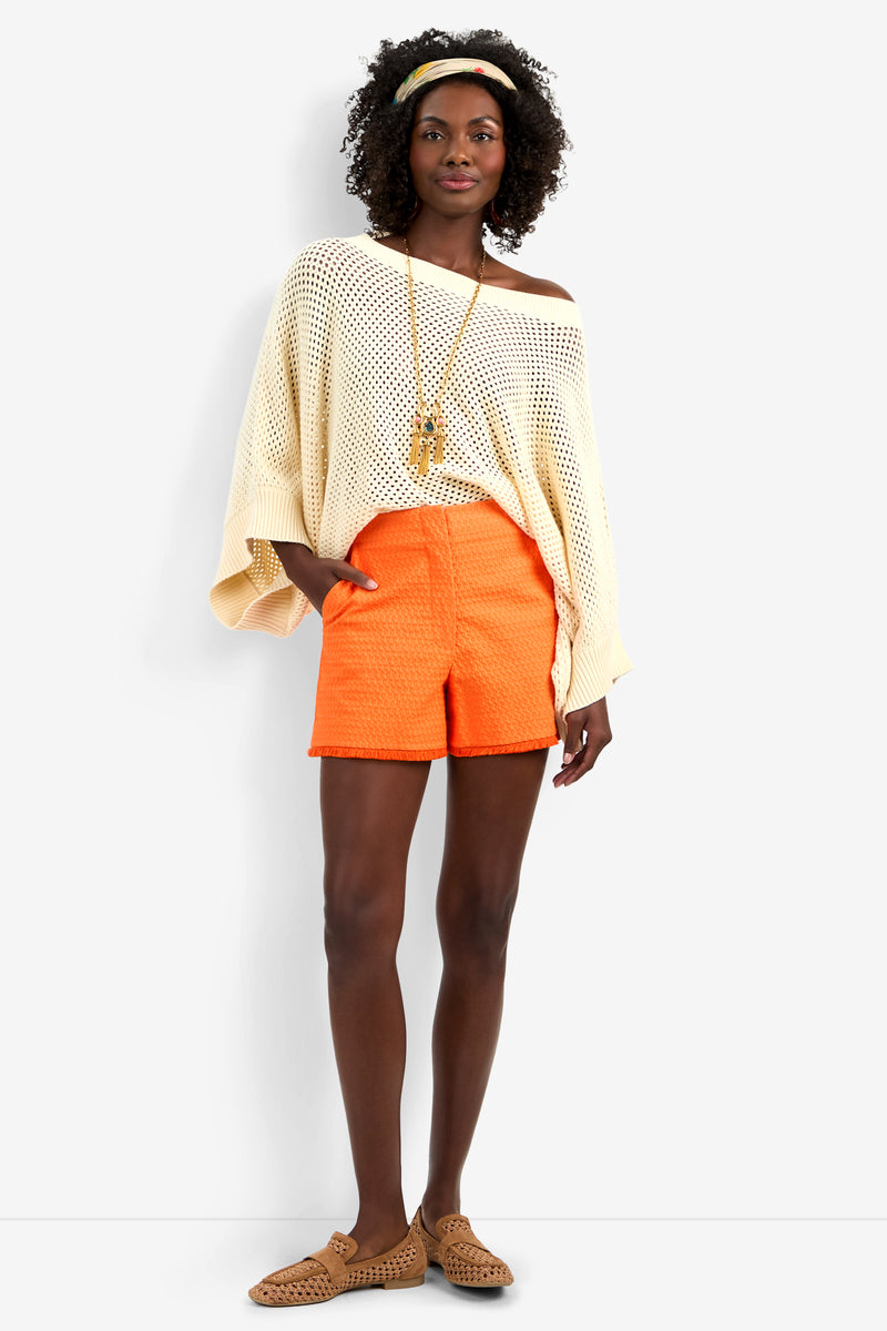 A woman stands smiling against a plain background, wearing the Horizon Pullover—a loose cream knit top—with orange shorts, tan woven sandals, a long necklace, and a yellow headband, one hand tucked in her pocket.