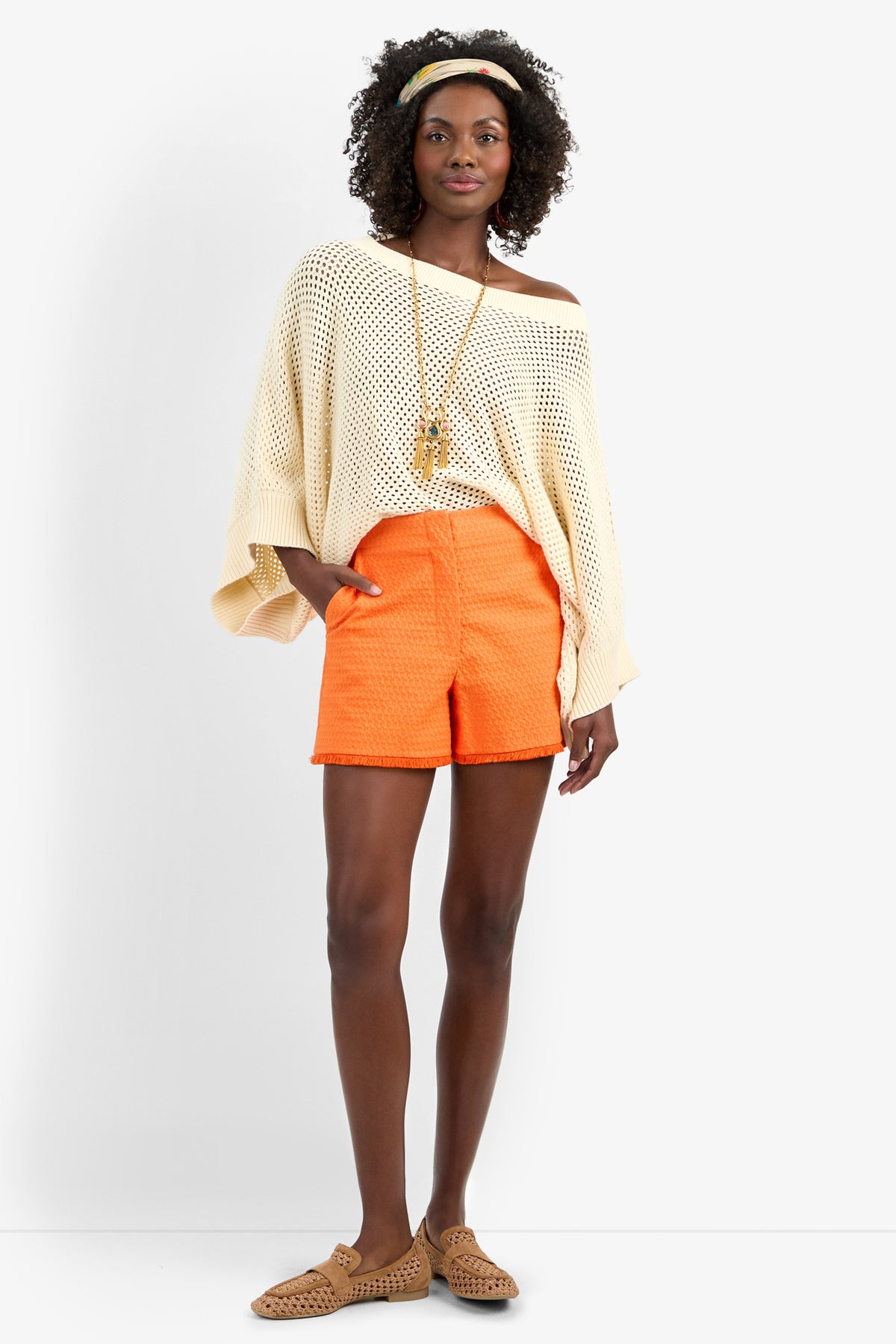 A woman stands against a plain background wearing the HORIZON PULLOVER, an off-the-shoulder cream knit sweater, paired with orange textured cotton shorts, tan woven shoes, a long necklace, and a headband.