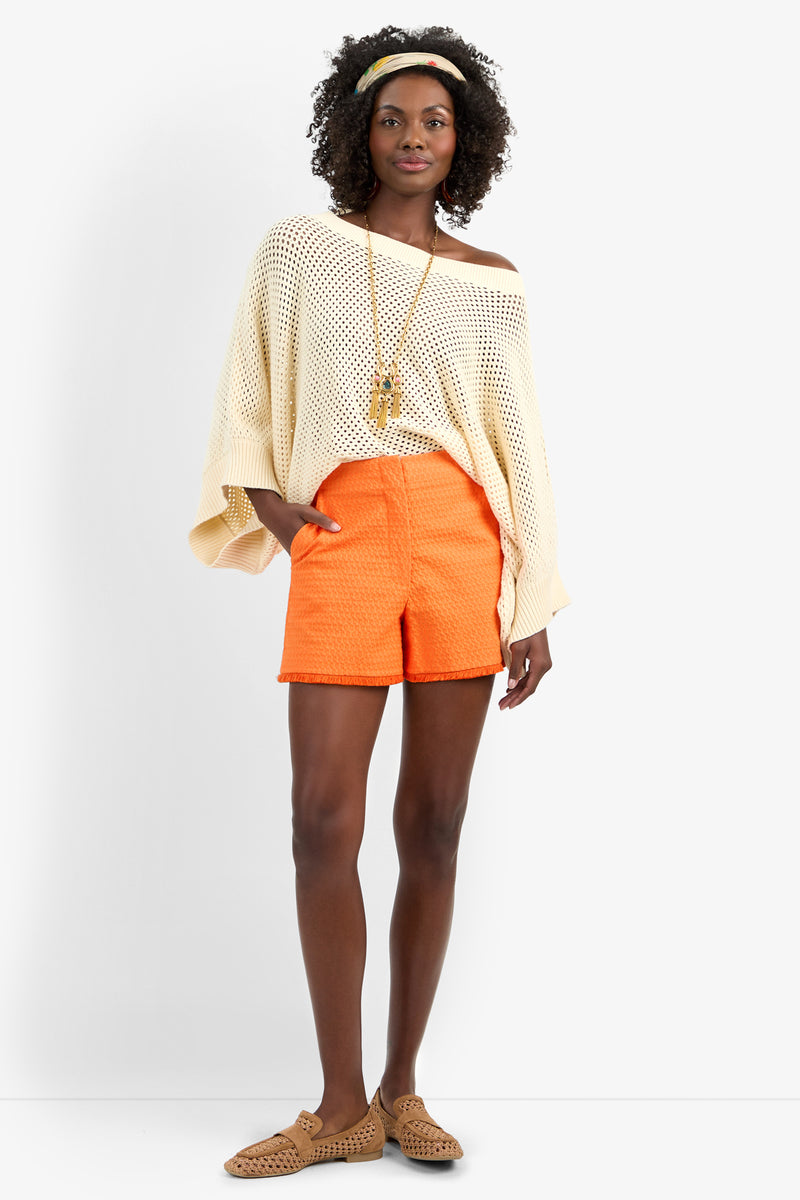 A woman stands against a plain background wearing the HORIZON PULLOVER, an off-the-shoulder cream knit sweater, paired with orange textured cotton shorts, tan woven shoes, a long necklace, and a headband.
