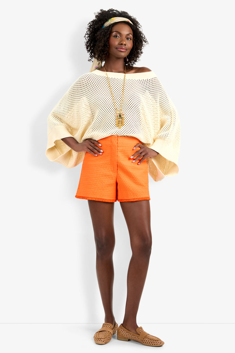 A woman stands confidently with hands on hips against a plain background, wearing the Horizon Pullover—a loose cream knit top—paired with bright orange shorts, tan slip-on shoes, a headband, and a long necklace.