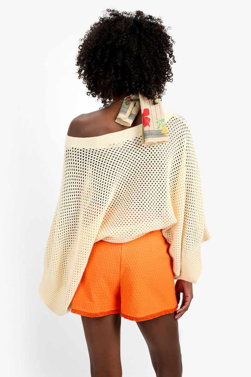 A person stands facing away, wearing the HORIZON PULLOVER—an off-the-shoulder cream knit cotton sweater—with bright orange textured shorts and a patterned scarf in curly hair, set against a plain white background.