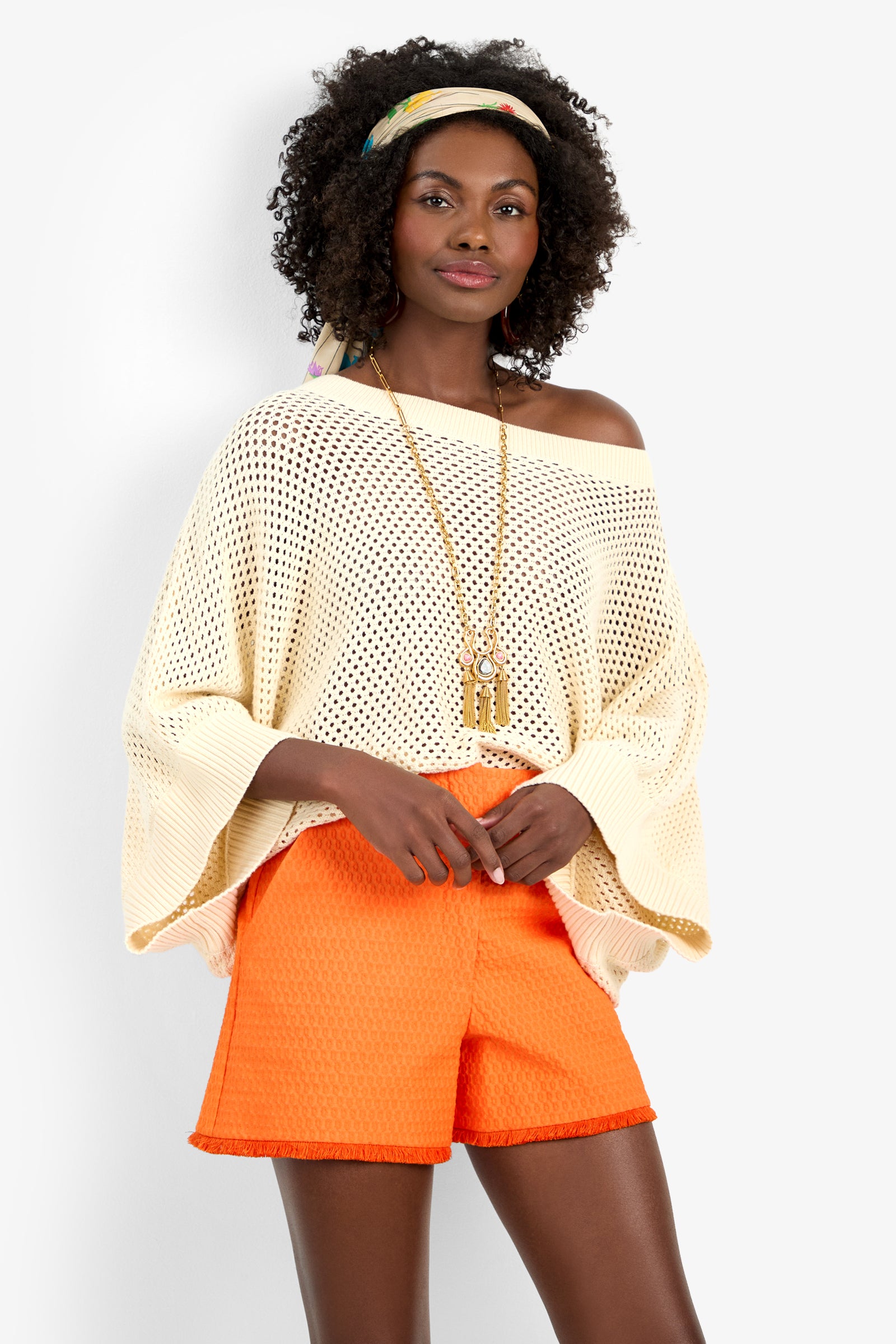 A woman stands confidently against a plain white background, wearing the HORIZON PULLOVER off-shoulder cream knit sweater, bright orange textured cotton shorts, a long gold necklace, and a colorful headscarf.