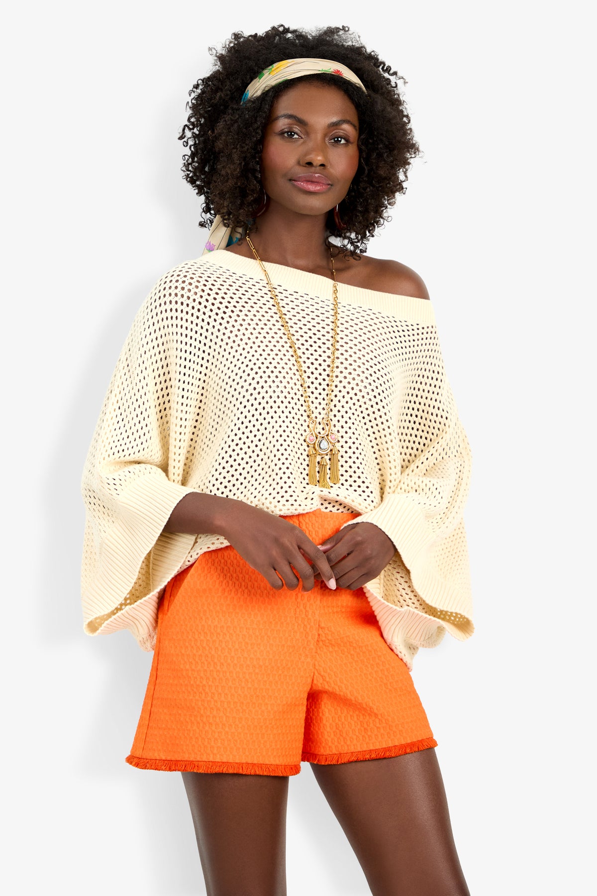 Wearing the off-white Horizon Pullover and bold orange shorts, she stands confidently with hands in pockets, complemented by a colorful headscarf, statement earrings, and a long necklace.