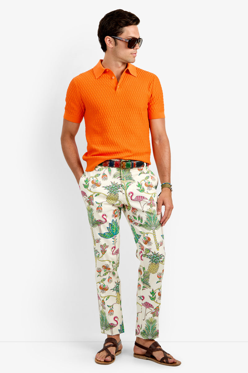 A man in sunglasses, the HONALO SS POLO (orange, 100% cotton), white pants with tropical animal prints, a multicolored belt, and brown sandals stands against a plain white background.