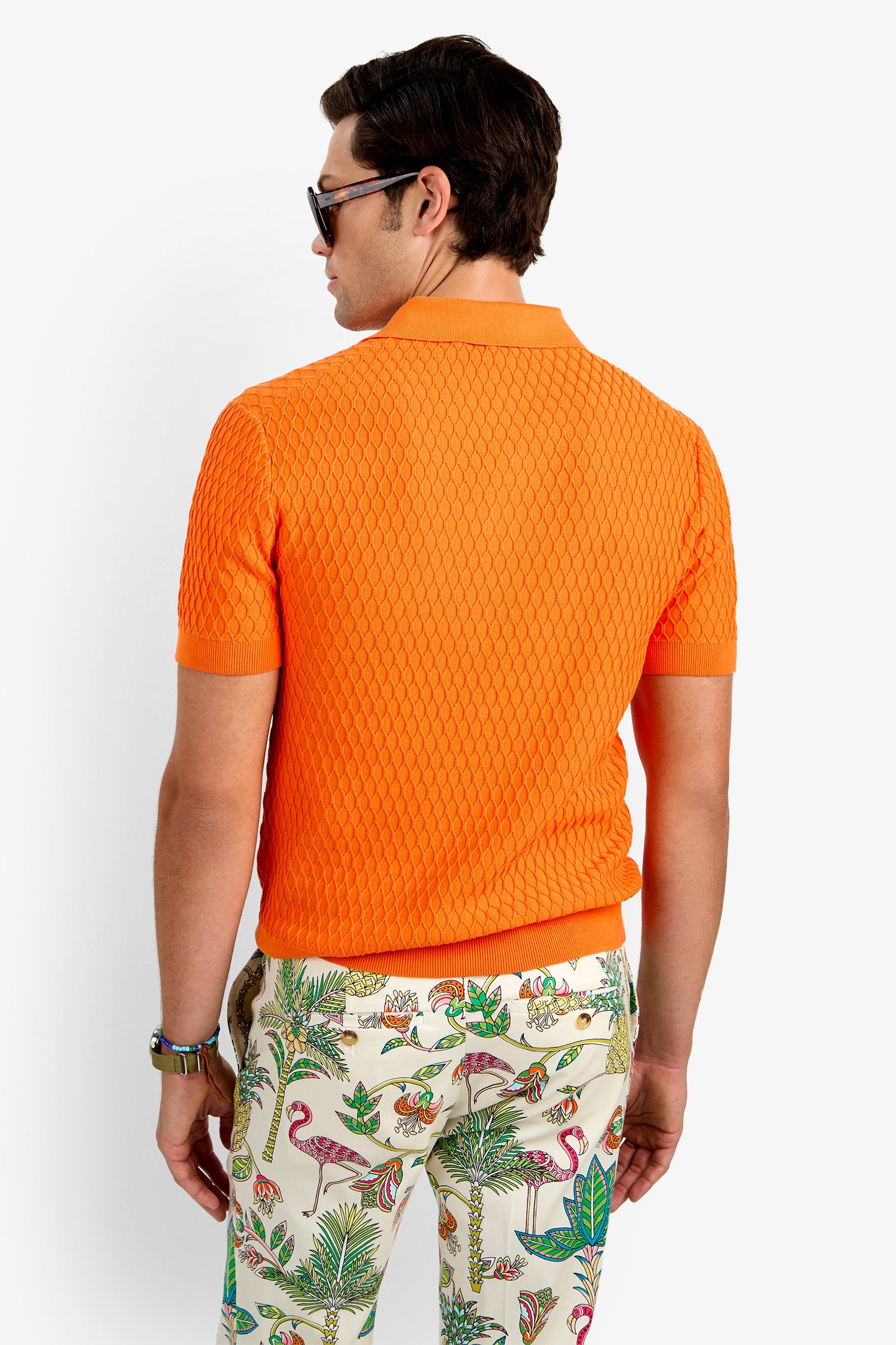 A man with dark hair in sunglasses, wearing the HONALO SS POLO (a bright orange 100% cotton textured knit short-sleeve shirt) and patterned pants with colorful animals and plants, stands facing away from the camera against a white background.