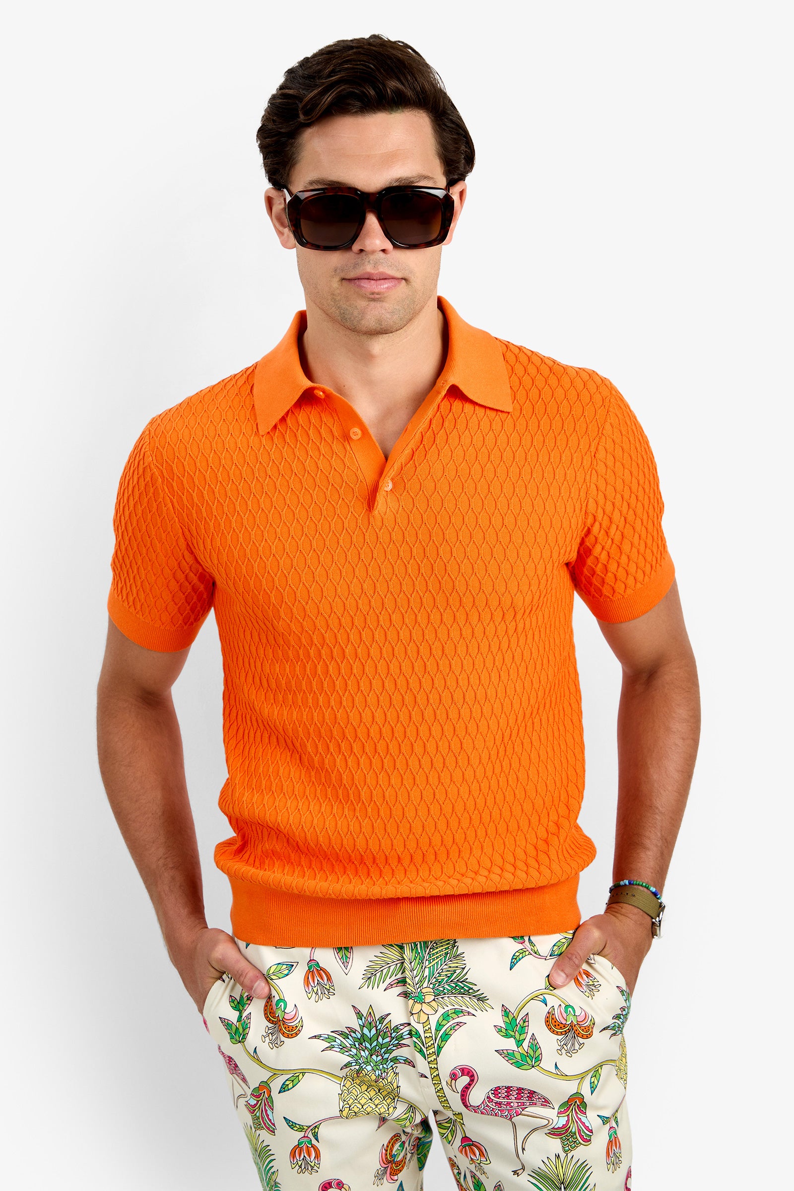 A man in large dark sunglasses and the HONALO SS POLO, a textured orange 100% cotton short-sleeve shirt, pairs it with white pants featuring colorful tropical prints, standing hands-in-pockets against a plain background.