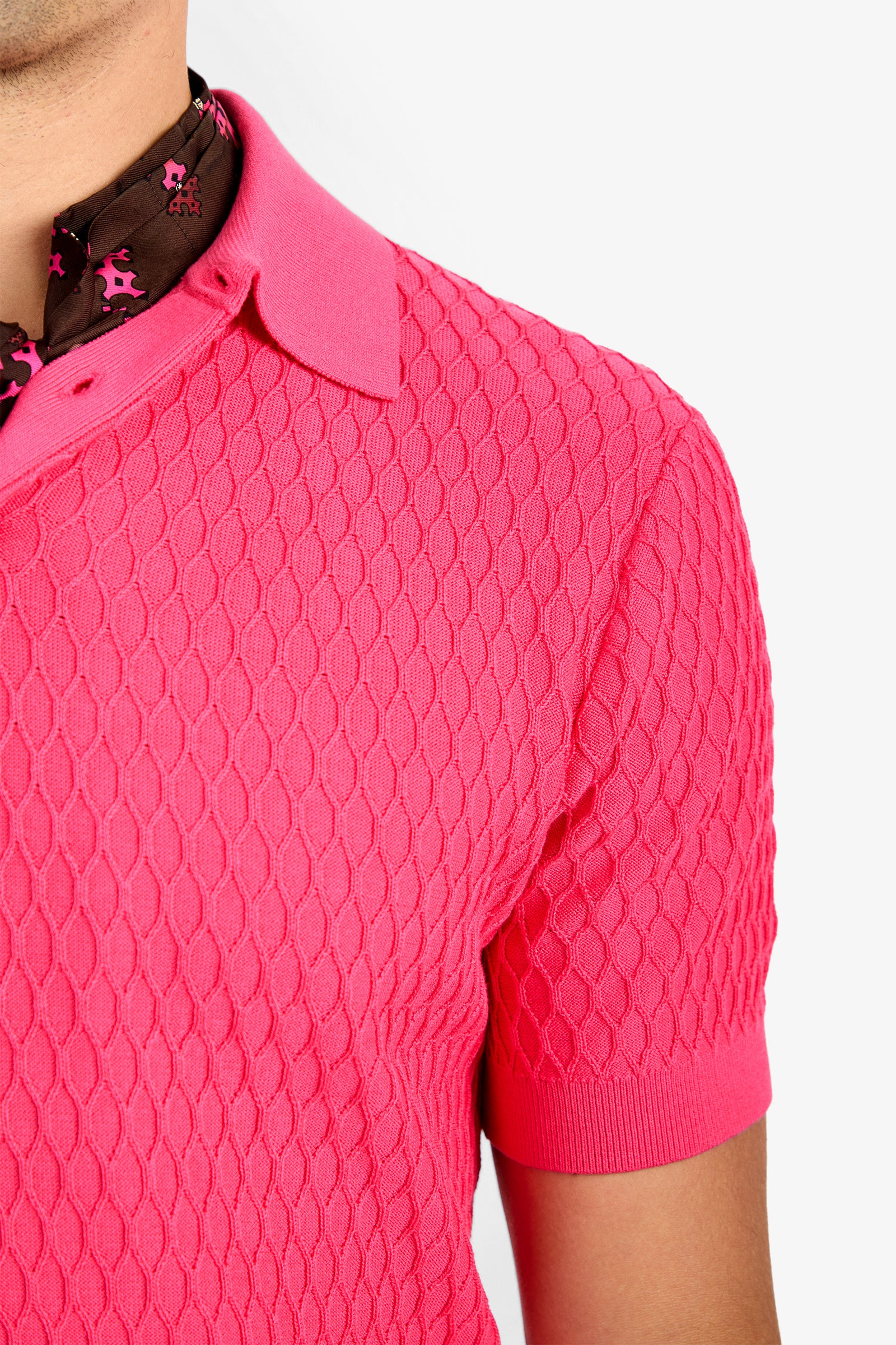 Close-up of a person wearing the HONALO SS POLO, a bright pink textured short-sleeve 100% cotton shirt, layered over a brown floral-patterned shirt, shown from the shoulders up against a white background.
