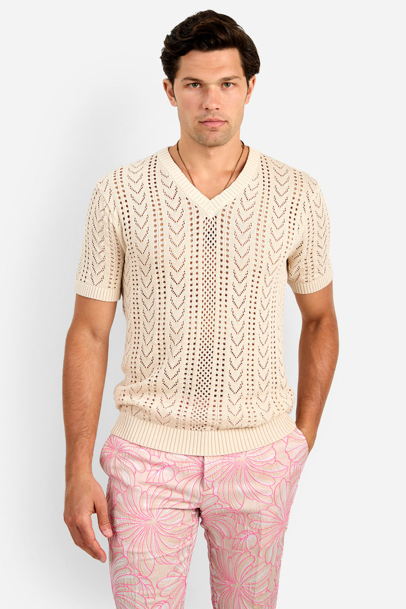 A man wears the KEANU CROCHET V-NECK, a cream short-sleeve cotton knit sweater with an openwork chevron pattern, paired with light pink floral pants, standing against a plain white background.