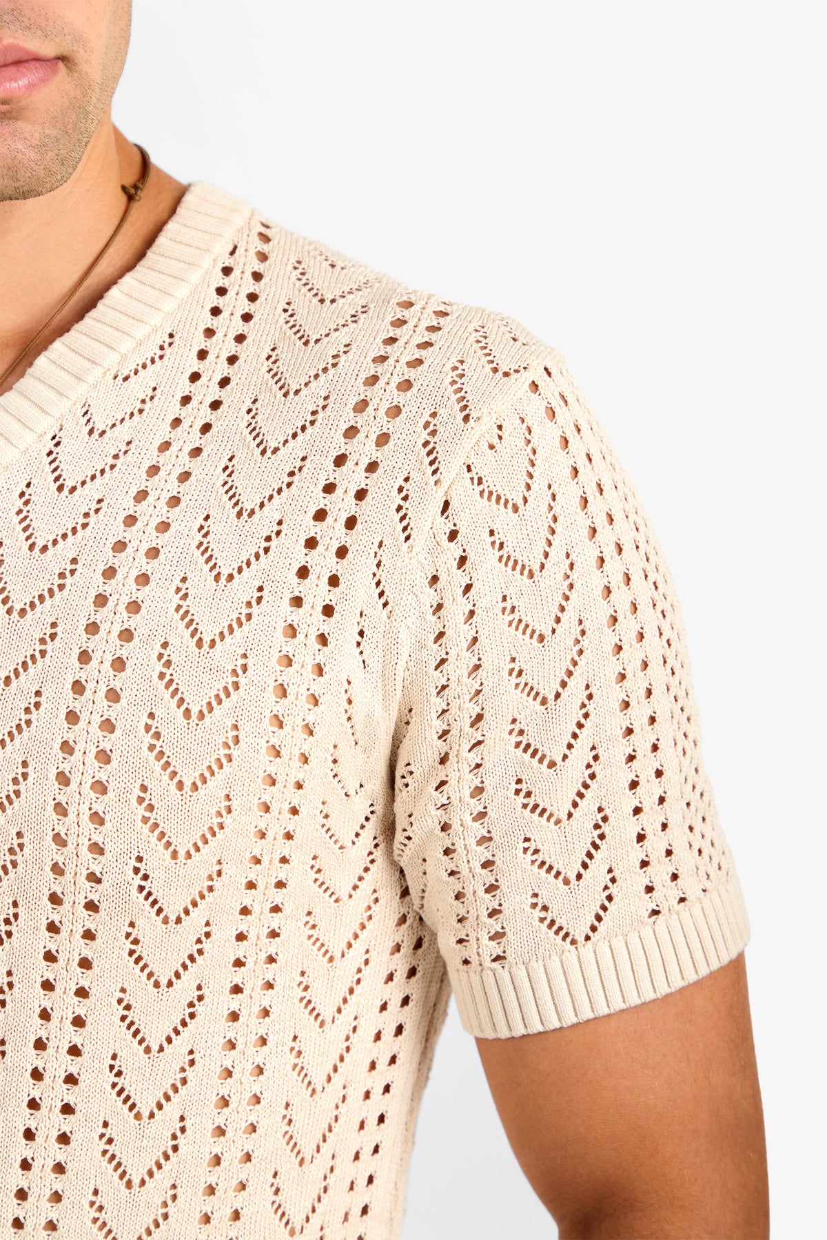 A close-up of a person wearing the KEANU CROCHET V-NECK, a beige short-sleeve knit shirt with chevron and eyelet patterns, ribbed collar, and sleeve cuffs. Only the torso, shoulder, and part of their face are visible.