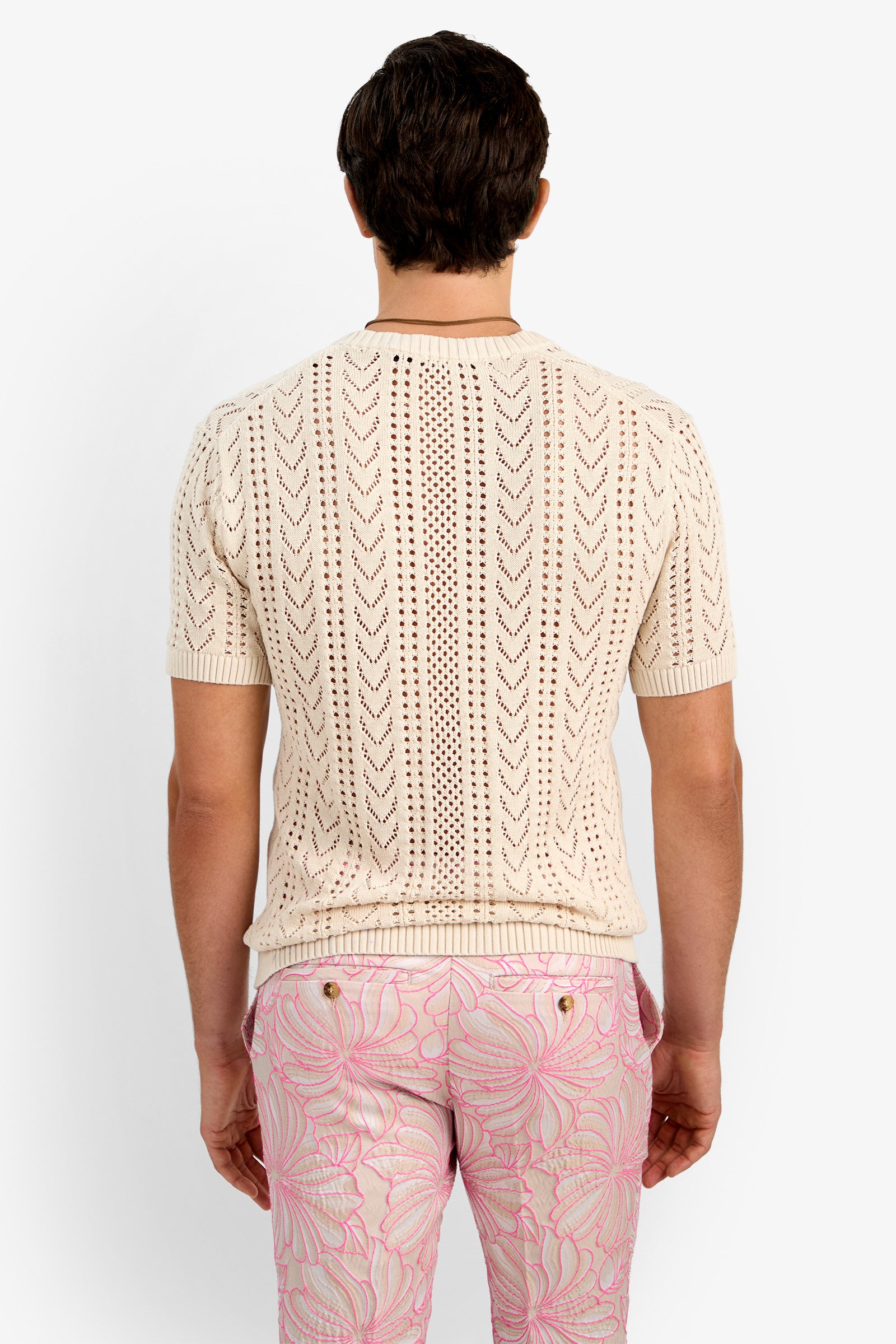 A man is seen from the back wearing the KEANU CROCHET V-NECK, a short-sleeve cream knit shirt with a chevron pattern, paired with light pink pants featuring a swirling abstract design.