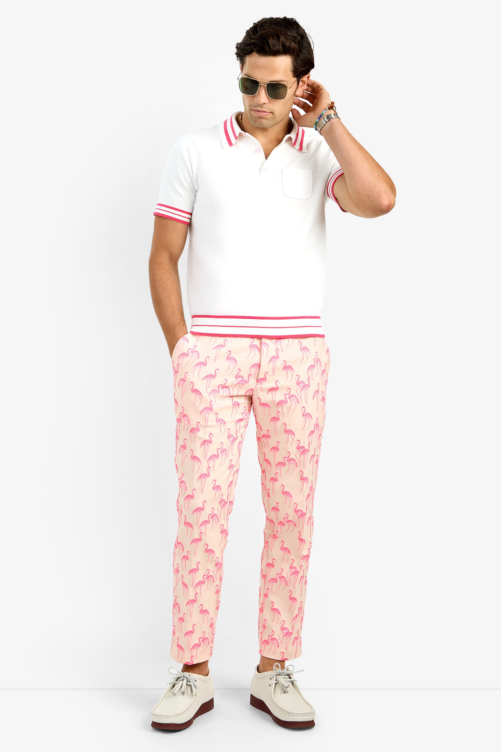 A man stands with his hands in his pockets, wearing sunglasses, a white polo shirt with a pink-striped collar and sleeves, and Clyde Slim Trousers featuring a light pink flamingo print, against a plain background.