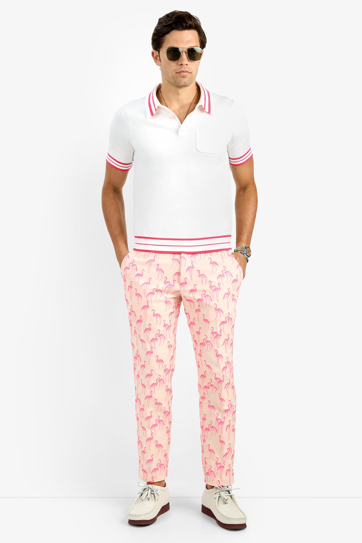 A man wearing sunglasses, a white polo shirt with pink trim, and Clyde Slim Trousers in light pink with a flamingo pattern stands with his hands in his pockets against a plain white background.