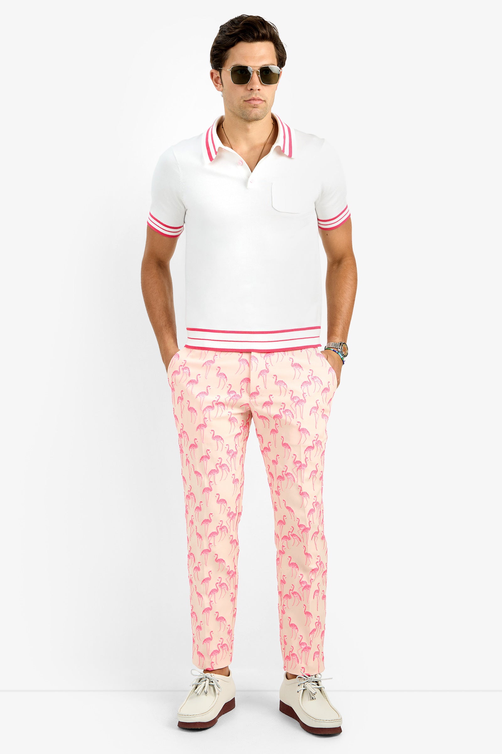 A man wearing sunglasses, a white polo shirt with pink trim, and Clyde Slim Trousers in light pink with a flamingo pattern stands with his hands in his pockets against a plain white background.