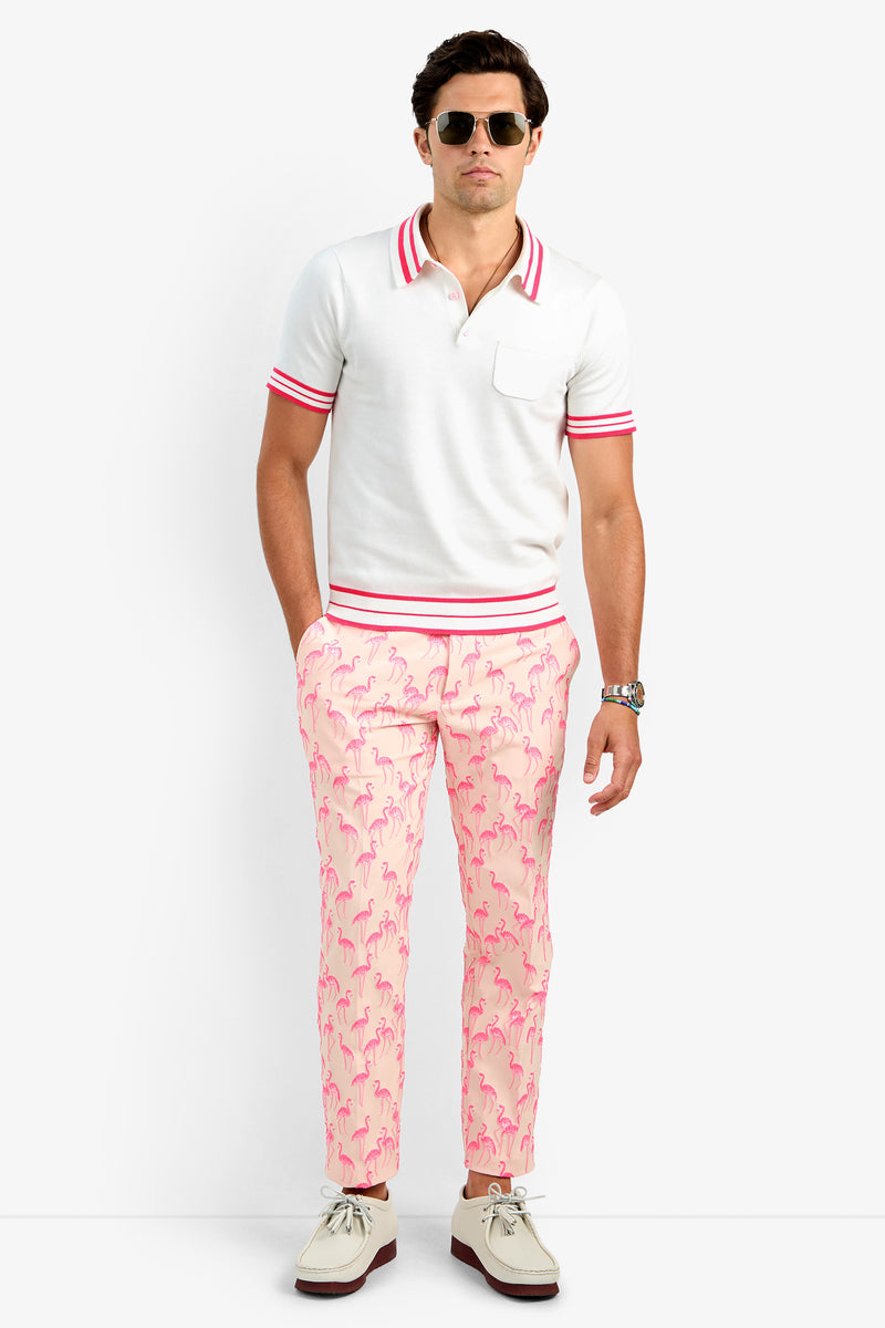 A man faces forward with his hands in his pockets, wearing sunglasses and the JAIME FLAMINGO POLO— a white 100% cotton polo shirt featuring red stripes on the collar and sleeves, paired with pink flamingo-patterned pants.