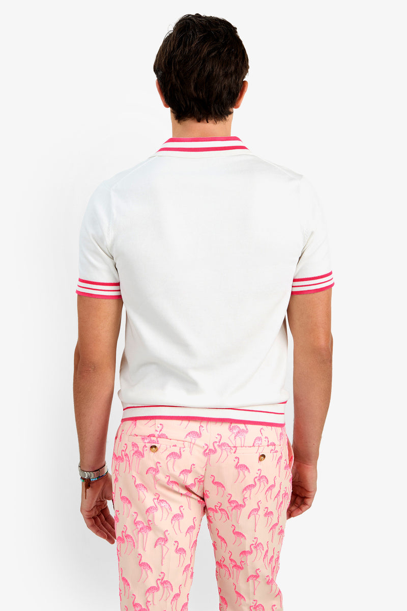 A man stands with his back to the camera, wearing the JAIME FLAMINGO POLO—a white cotton shirt with red striped trim—and pale pink flamingo-patterned pants.