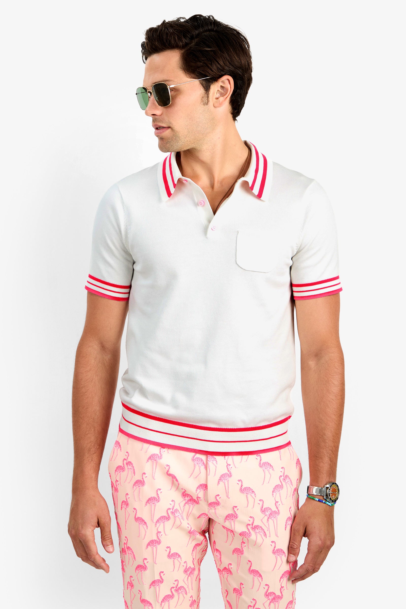 A man wearing aviator sunglasses and the JAIME FLAMINGO POLO in Whitewash, featuring red stripes on the collar, sleeves, and hem, paired with light pink flamingo-print pants, stands looking to his left.