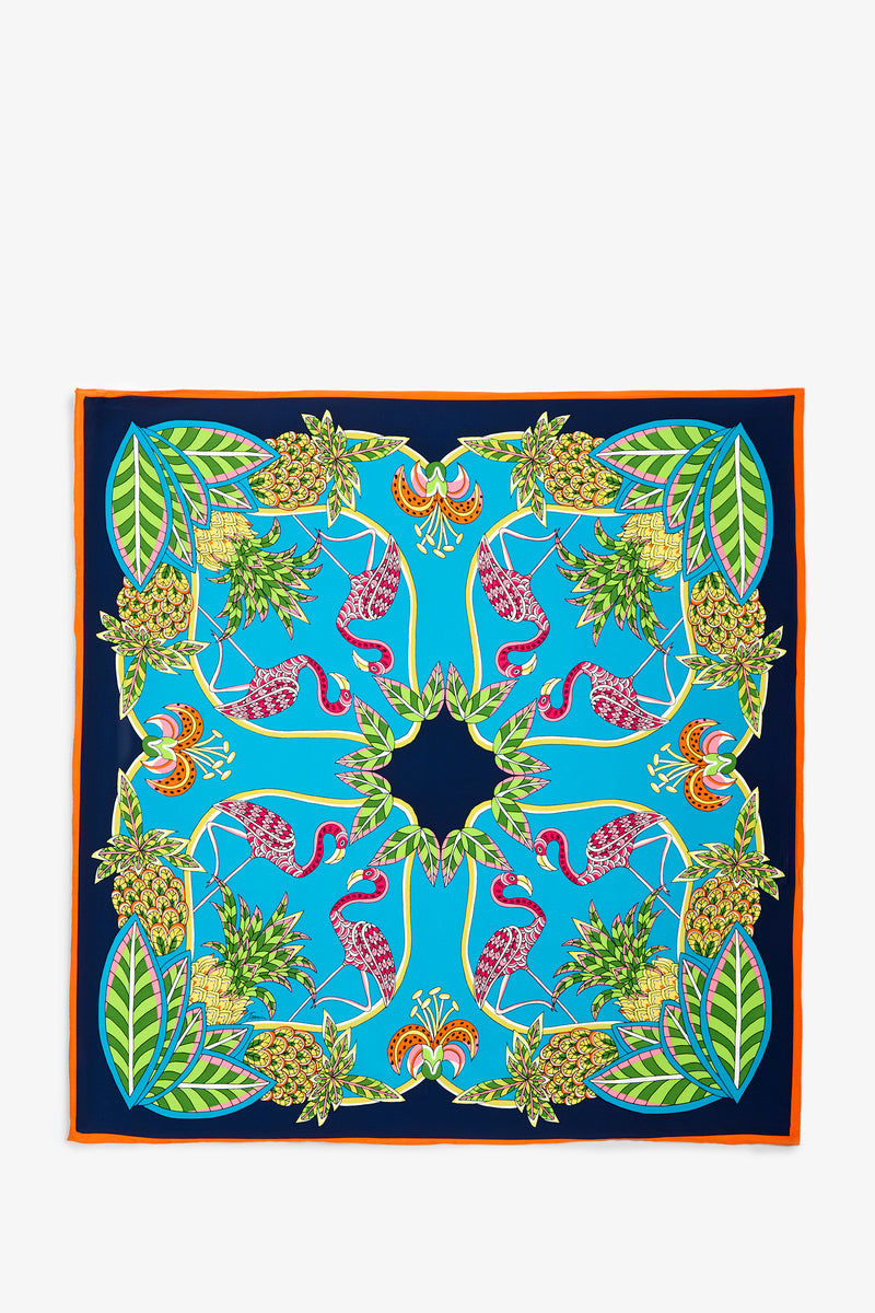 The Tiger Lily Scarf is a square scarf featuring a vibrant pattern of pink flamingos, green leaves, and pineapples on a blue background, finished with an orange border.