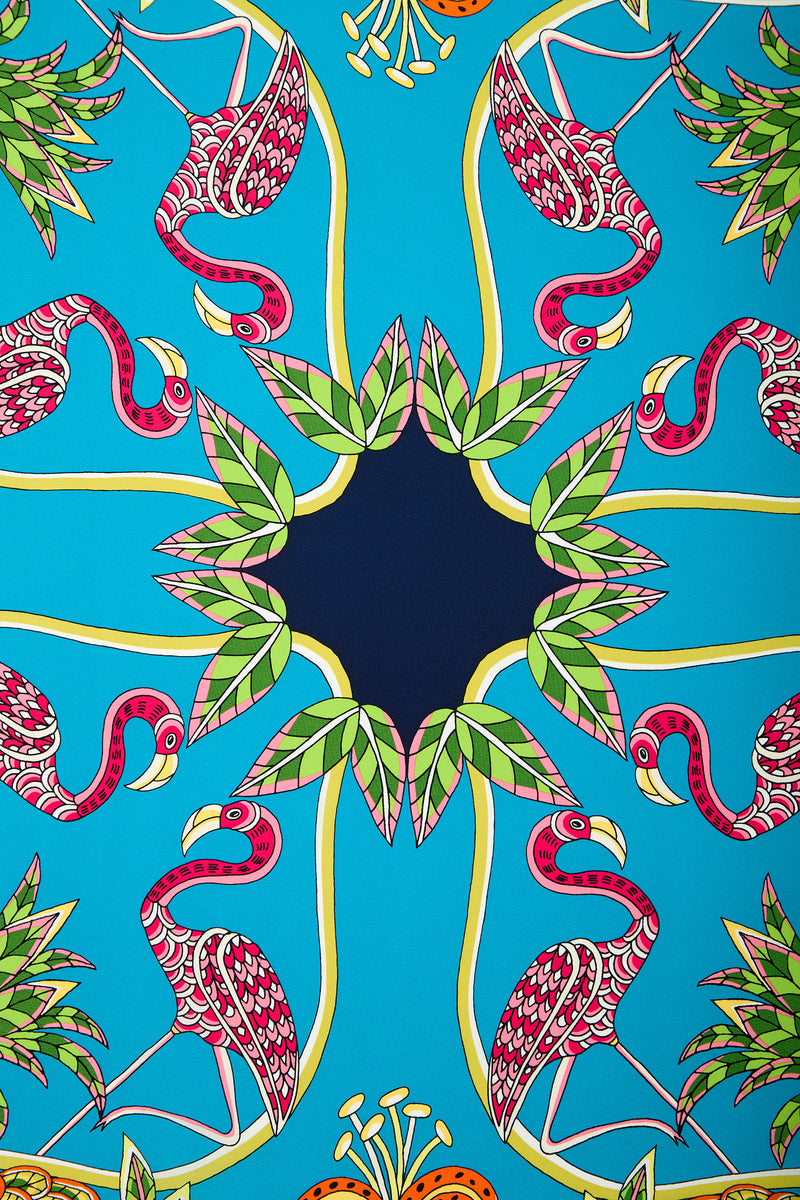 Colorful illustration featuring pink flamingos and green leaves arranged in a symmetrical, kaleidoscope-like pattern on a bright blue background, forming a circular design with a dark blue center—featured on the Tiger Lily Scarf.