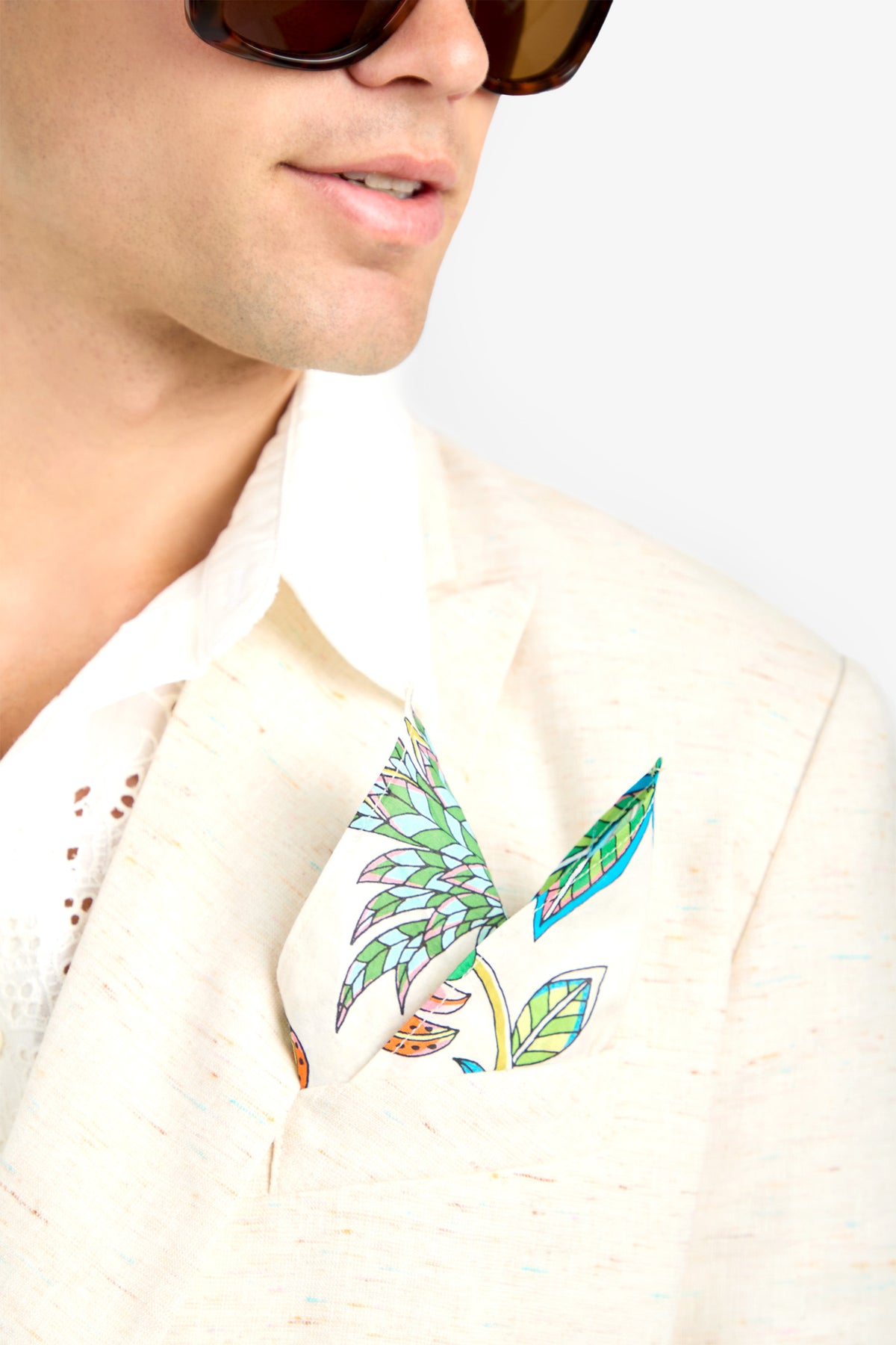 A man in a light beige, 100% cotton suit and white shirt wears sunglasses. A brightly colored PKT SQR floral-patterned pocket square is neatly folded in his jacket pocket. Only his lower face and upper torso are visible.
