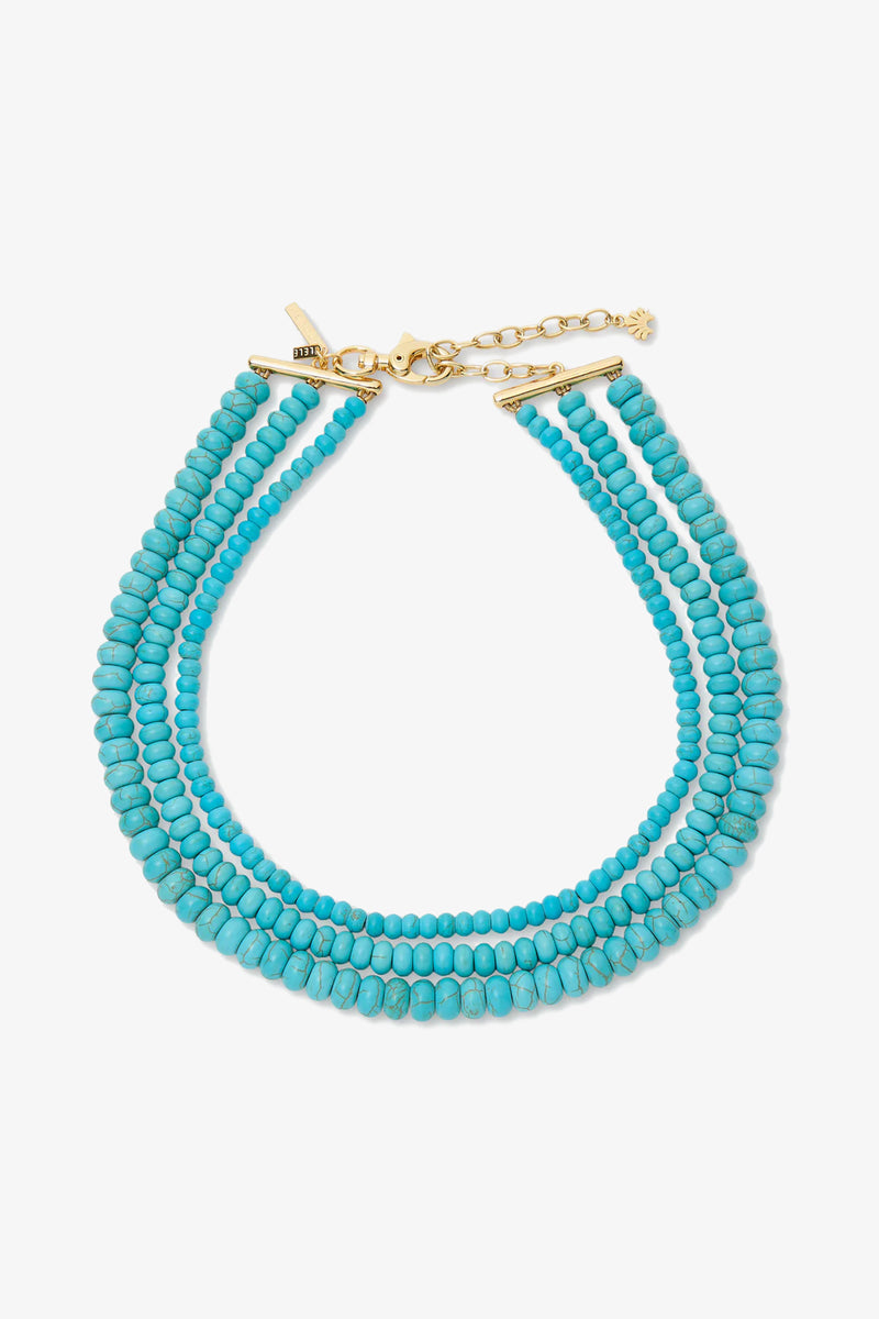 The Turquoise Multi Strand Necklace features three strands of small round turquoise beads with a gold clasp and extender chain, displayed on a white background.