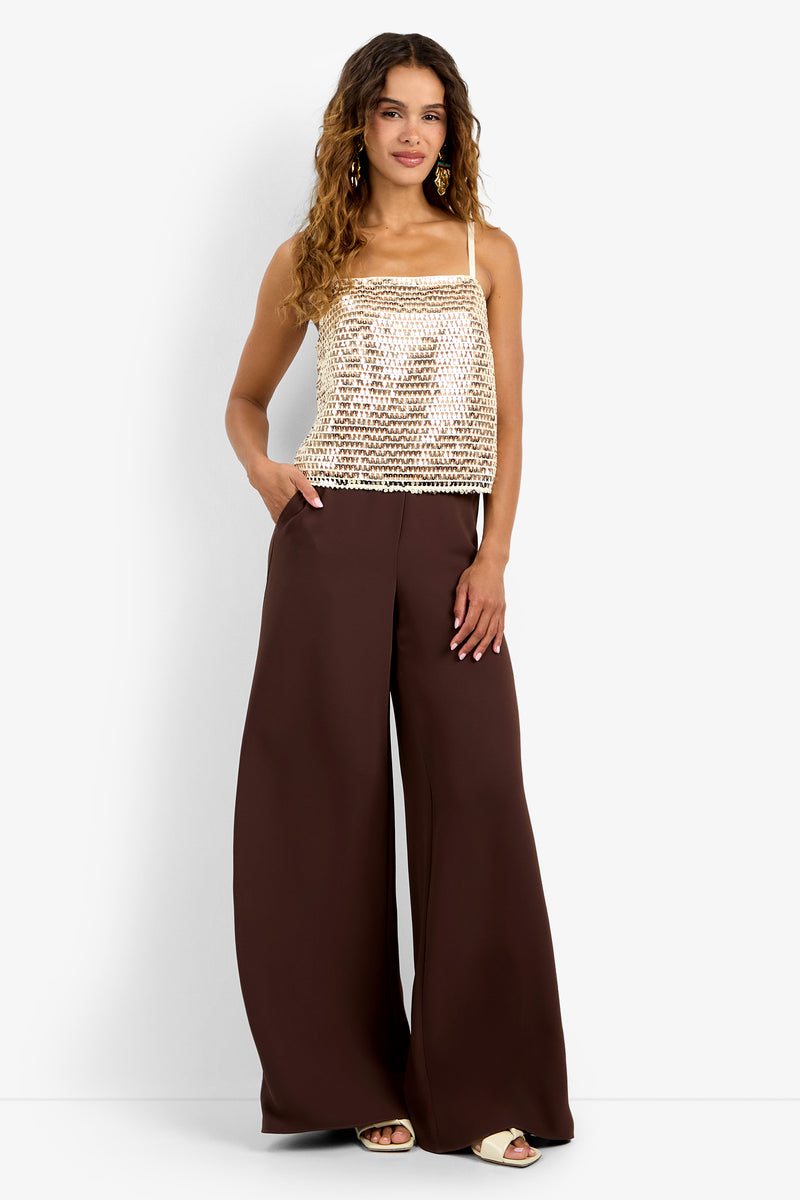 A woman wears the Alannie Top, a cream sequin tank, with high-waisted wide-leg brown pants. She has long curly hair, gold earrings, and beige sandals, posing with one hand in her pocket against a plain background.