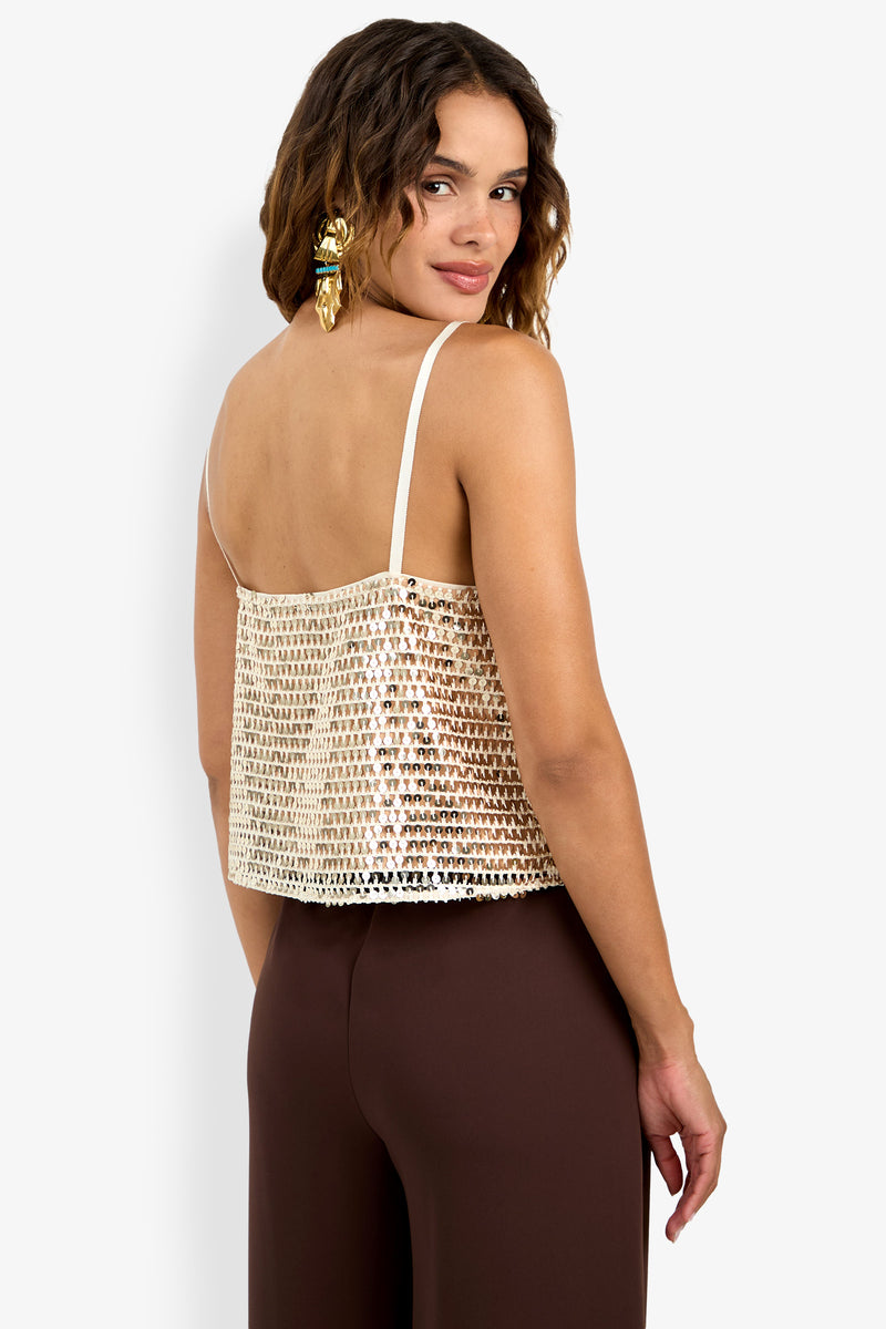 Woman with wavy brown hair and statement earrings poses with her back to the camera, wearing the Alannie Top—a gold sequin camisole with white straps—and brown high-waisted pants, glancing over her shoulder with a subtle smile.