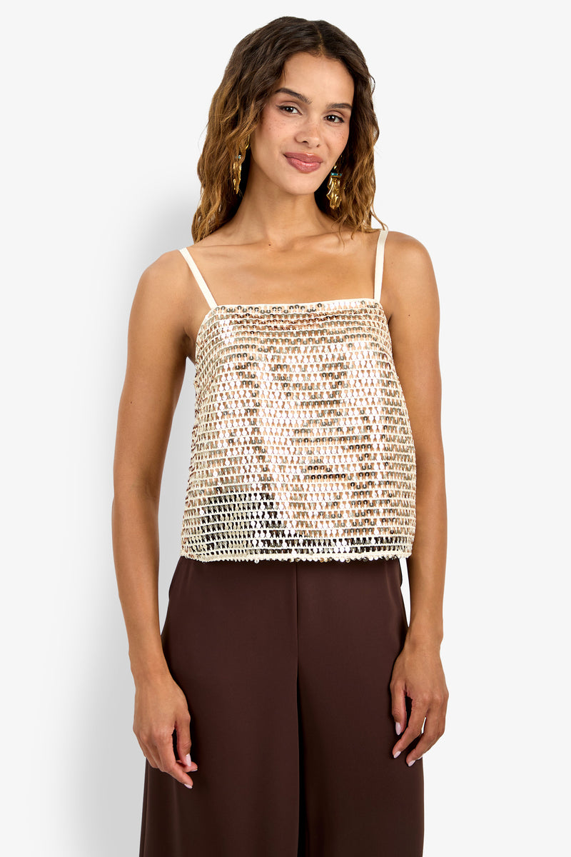 A woman with wavy brown hair wears the gold sequined Alannie Top with thin straps and brown wide-leg pants, standing against a plain white background.