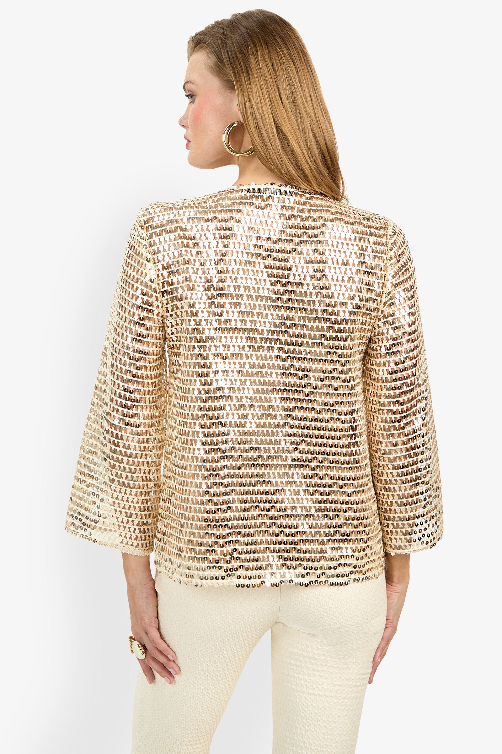 A woman with light brown hair is seen from behind wearing the Mailani Top, a shiny gold sequin long-sleeve jacket, paired with cream textured pants, hoop earrings, and a statement ring against a plain white background.