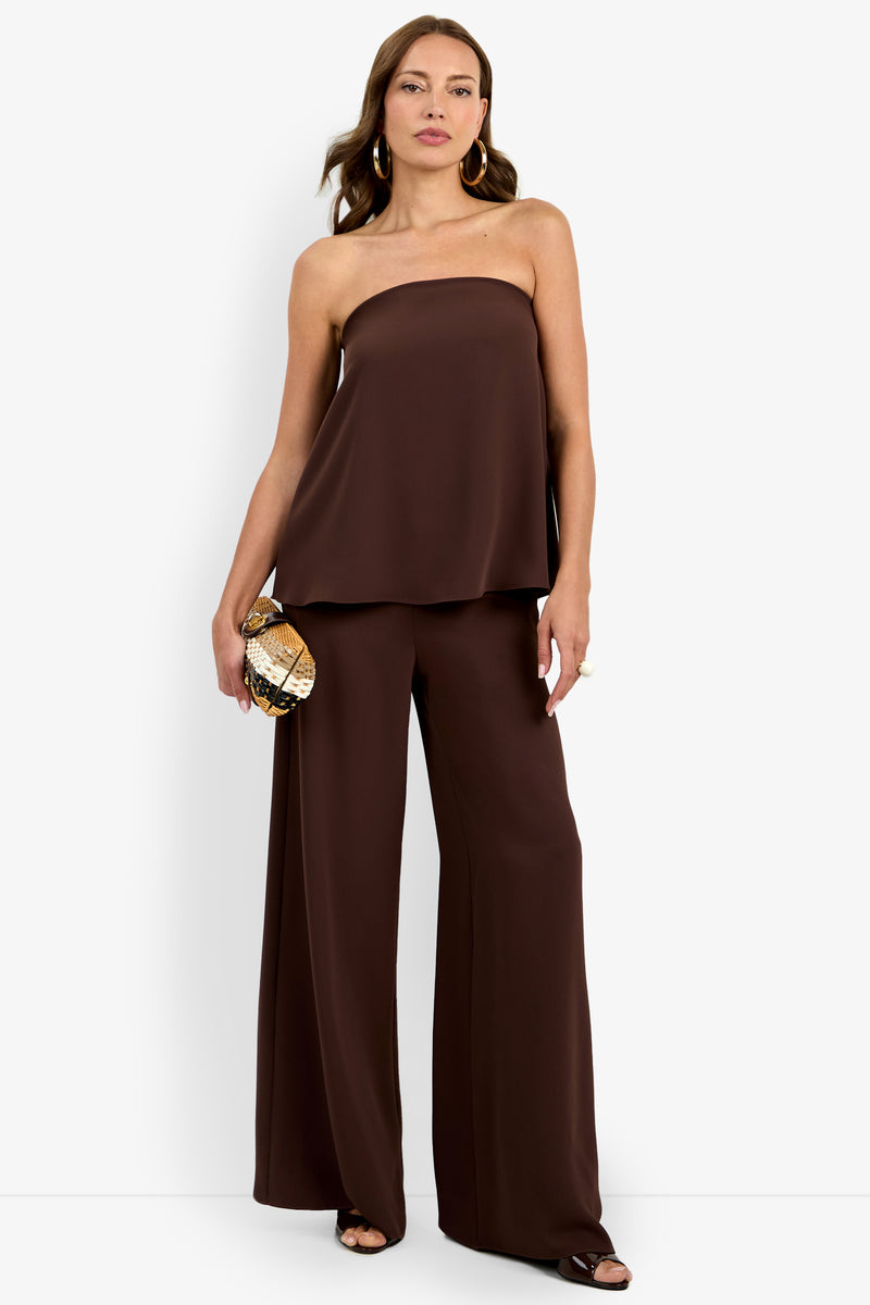 Woman wearing the Hulla Top, a strapless, loose-fitting dark brown top, paired with matching wide-leg pants. She holds a small patterned clutch and accessorizes with gold hoop earrings and a ring, standing against a plain background.