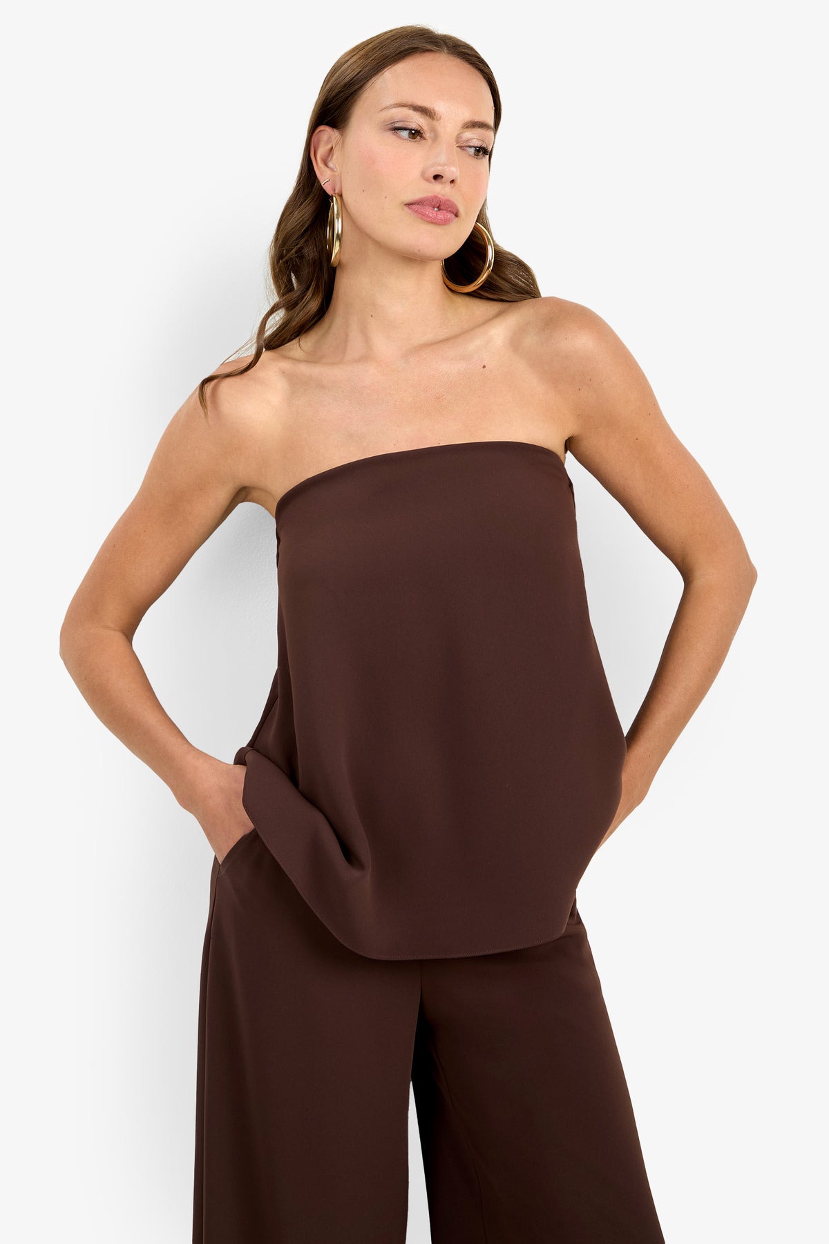 A woman with long brown hair and large hoop earrings stands against a white background, hands in her pockets, wearing the strapless dark brown Hulla Top and matching wide-leg pants.