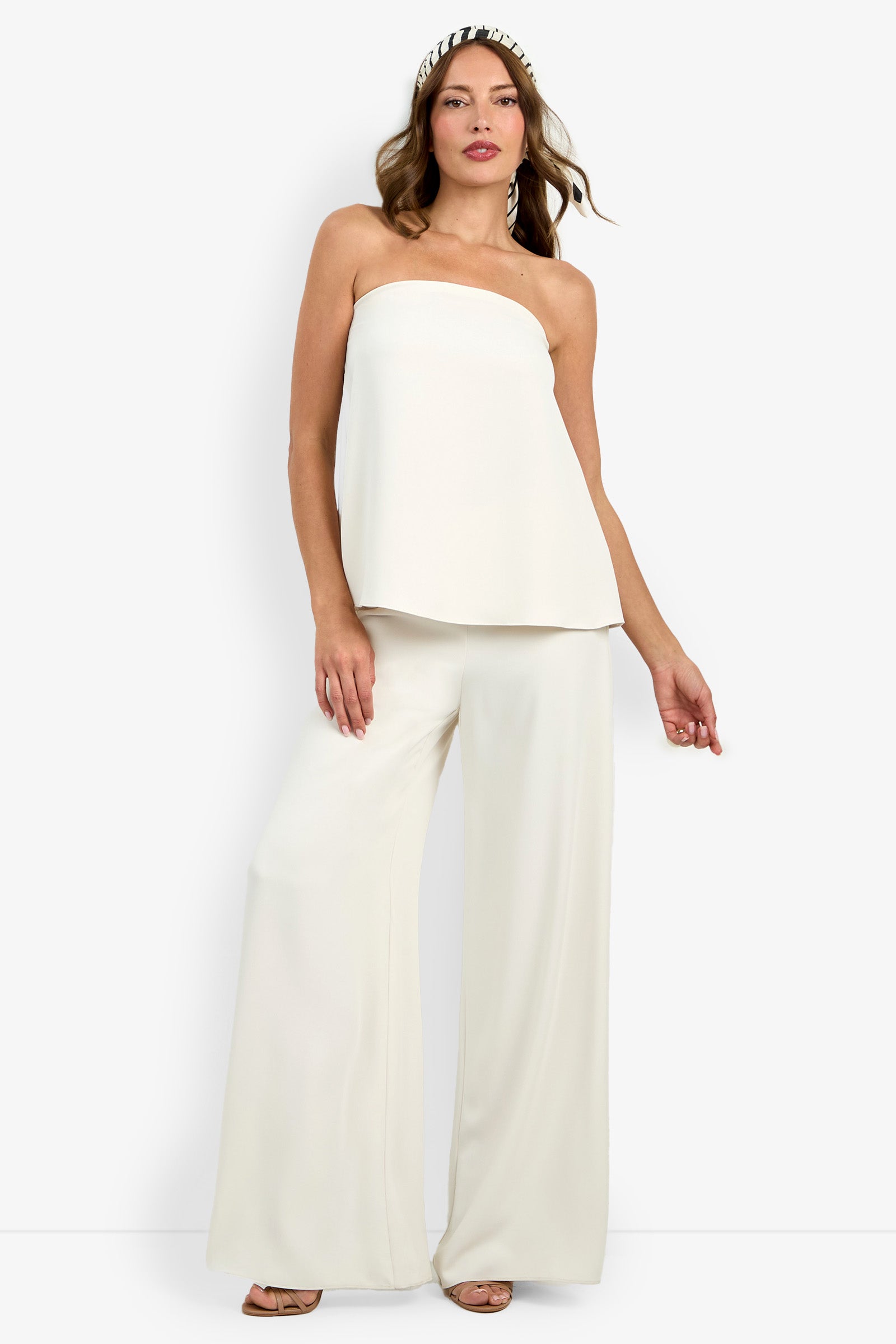 A woman wearing the Hulla Top, a strapless white top, pairs it with wide-leg white pants and accessorizes with a headband and open-toed heels against a white background.