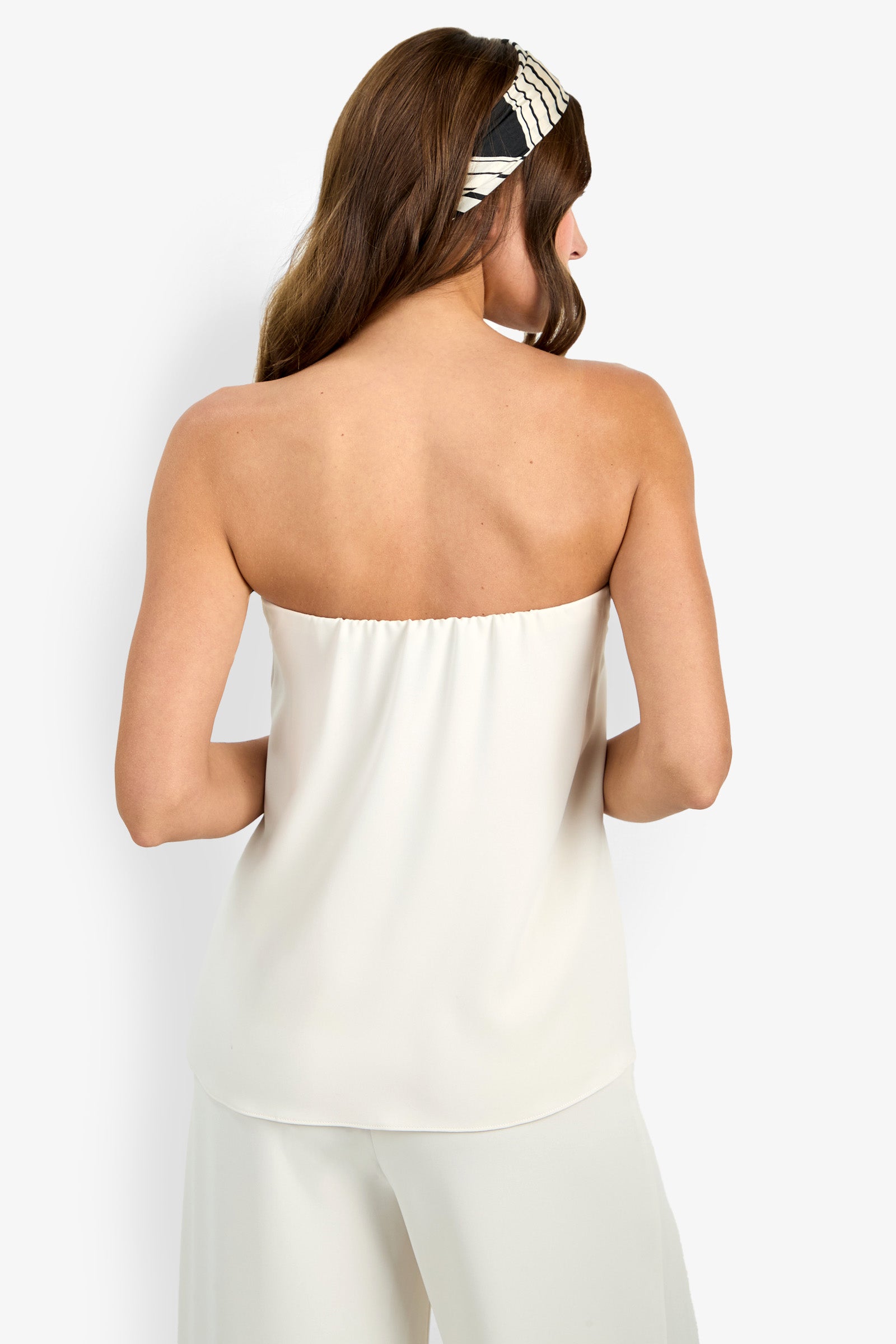 A woman with brown hair, seen from the back, models the Hulla Top—a white strapless top—paired with matching white pants and a black-and-white striped headband against a plain white background.
