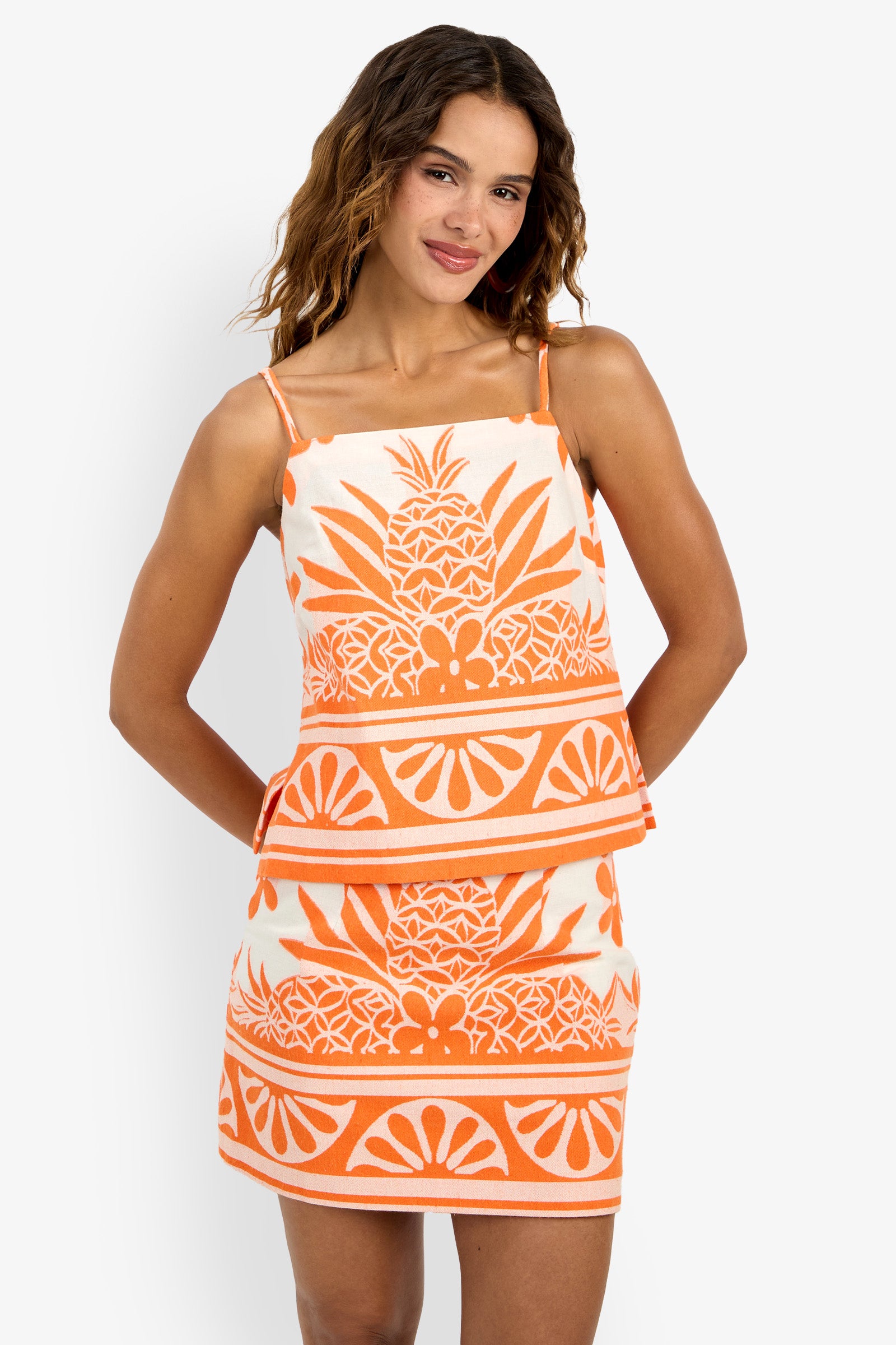A woman stands smiling with her hands behind her back against a white background, wearing the Marita Top—a sleeveless, orange and white piece featuring a tropical pineapple and geometric pattern.