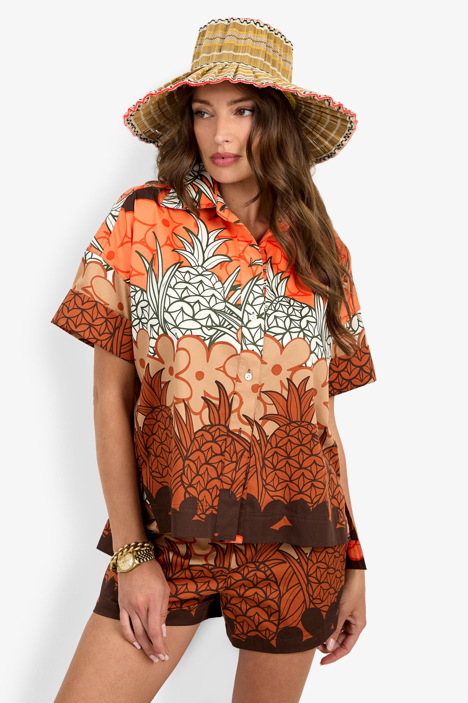 A woman wearing the Opponent Top, a short-sleeve button-up shirt with an orange and brown bold tropical pineapple print, stands against a plain white background.