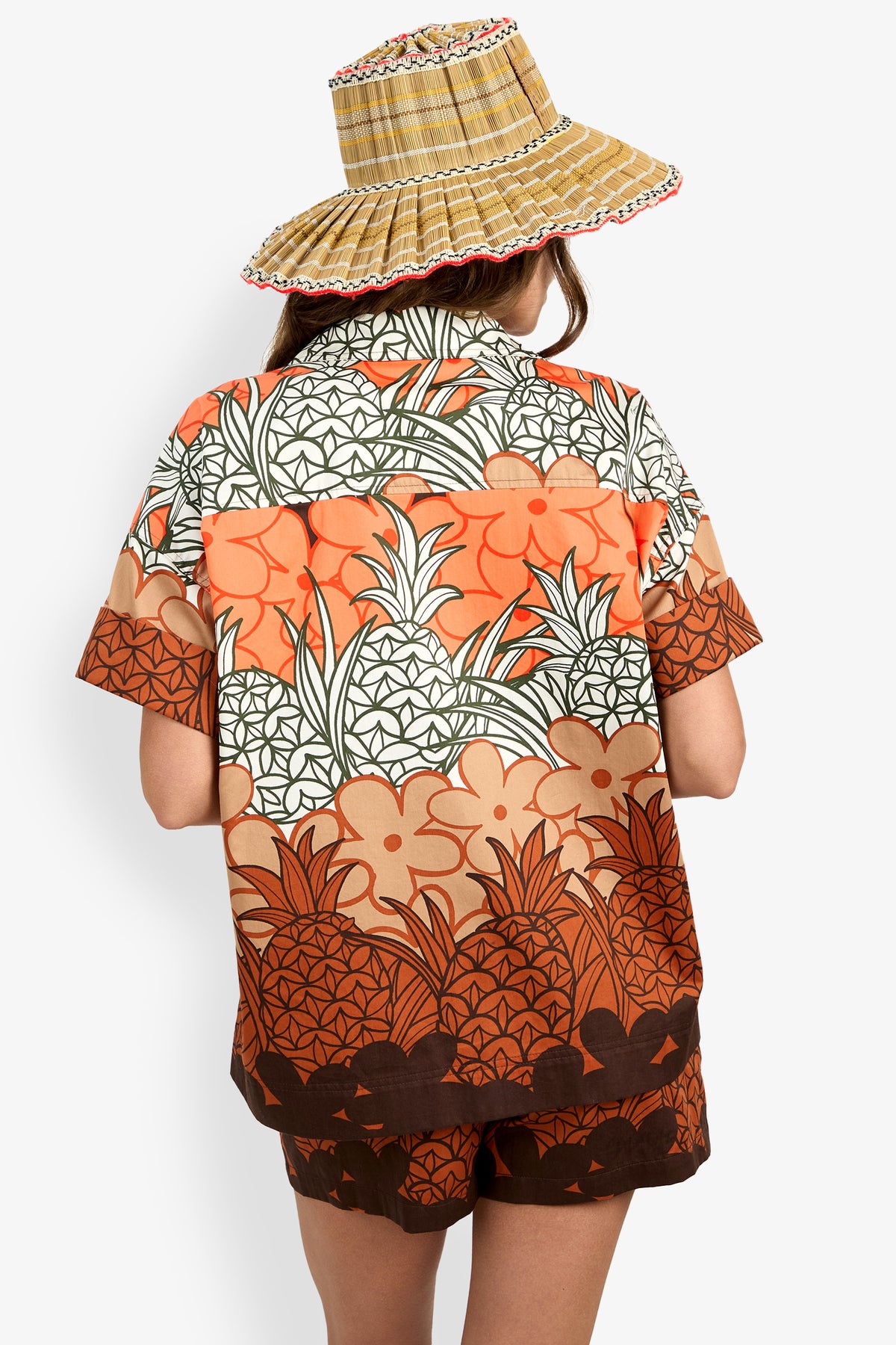A person is seen from behind wearing the Opponent Top, a short-sleeve shirt with an orange, brown, and white floral and geometric pattern, paired with matching shorts and a straw hat against a plain background.