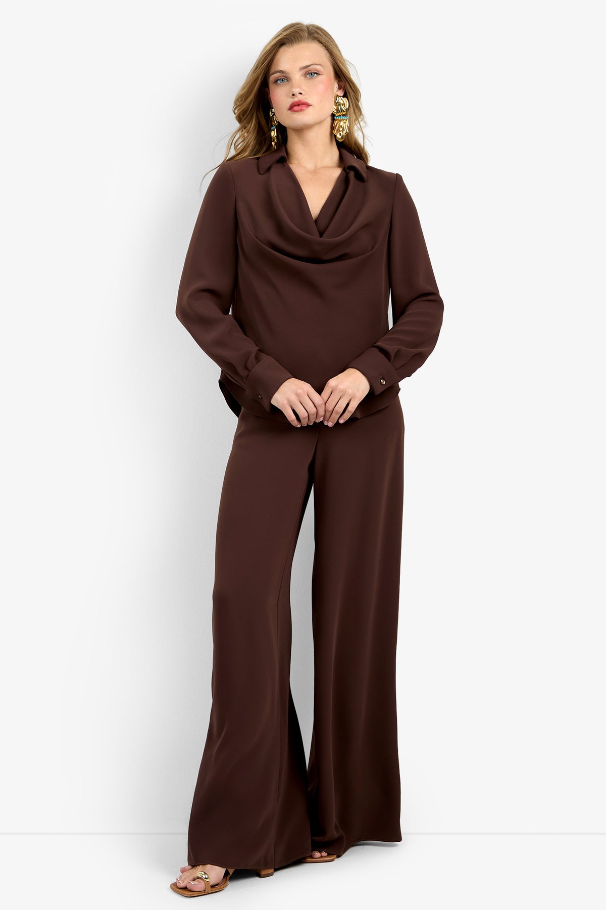 A woman poses against a white background in the MISTIC TOP in CAROB, a dark brown long-sleeve cowl-neck blouse with wide-leg matching pants, styled with gold earrings and tan sandals.