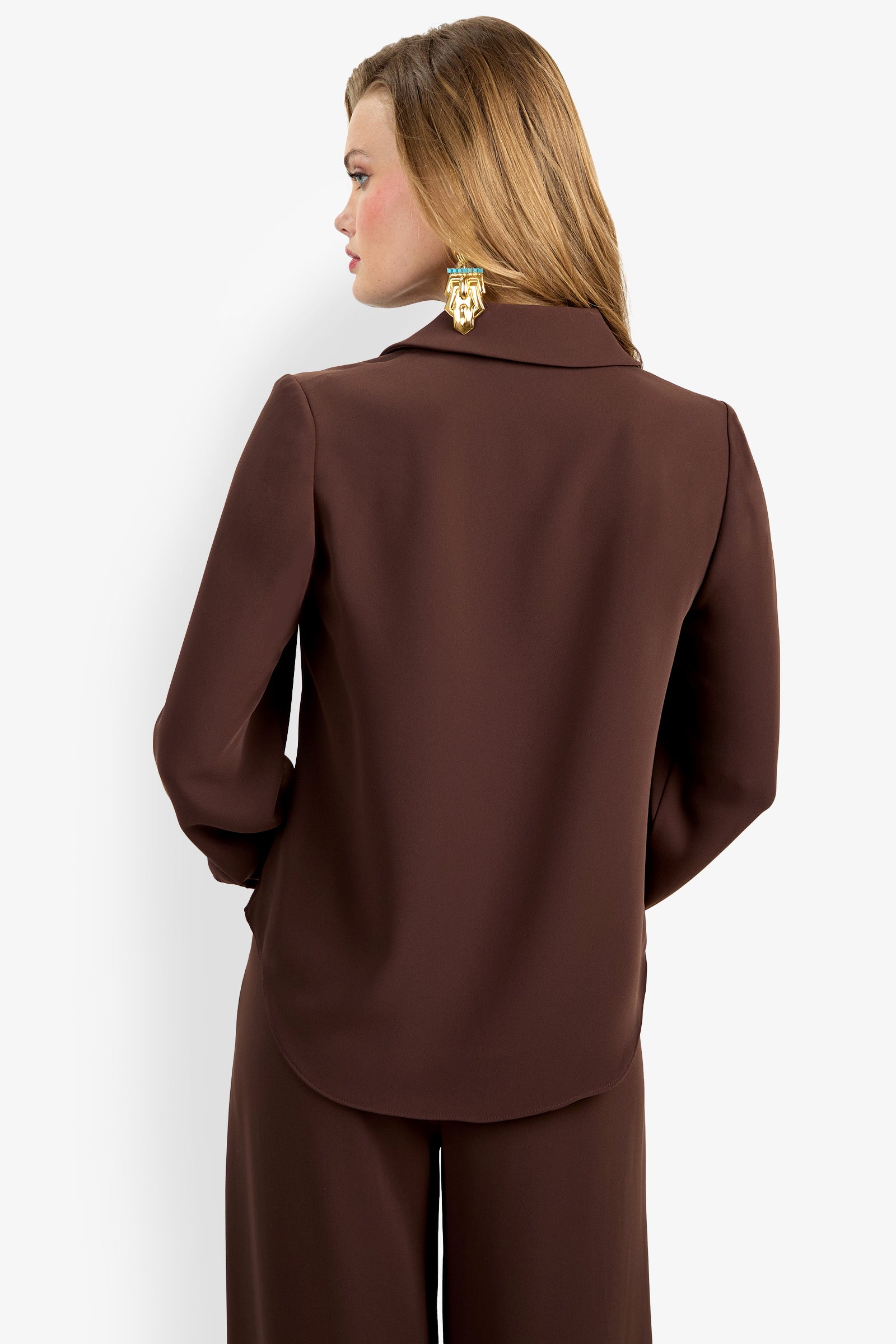 A woman with light brown hair is seen from the back wearing the Mistic Top, a dark brown long-sleeve blazer with matching pants. She has her left hand in her pocket, large gold earrings on, and stands against a white background.