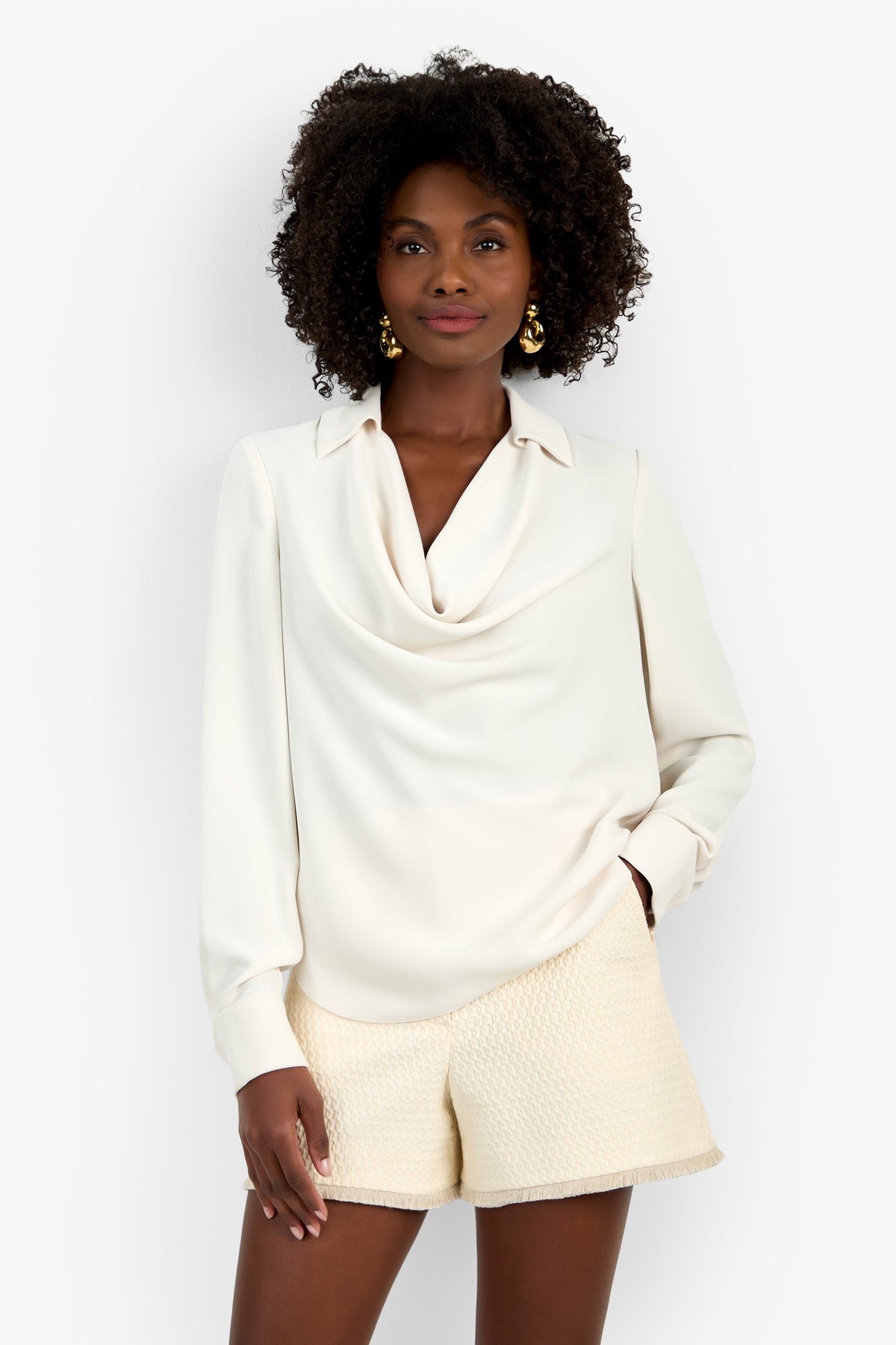 A woman stands against a white background wearing the cream Mistic Top with textured cream shorts, one hand in her pocket. She has curly hair, is smiling slightly, and accessorizes with gold earrings.