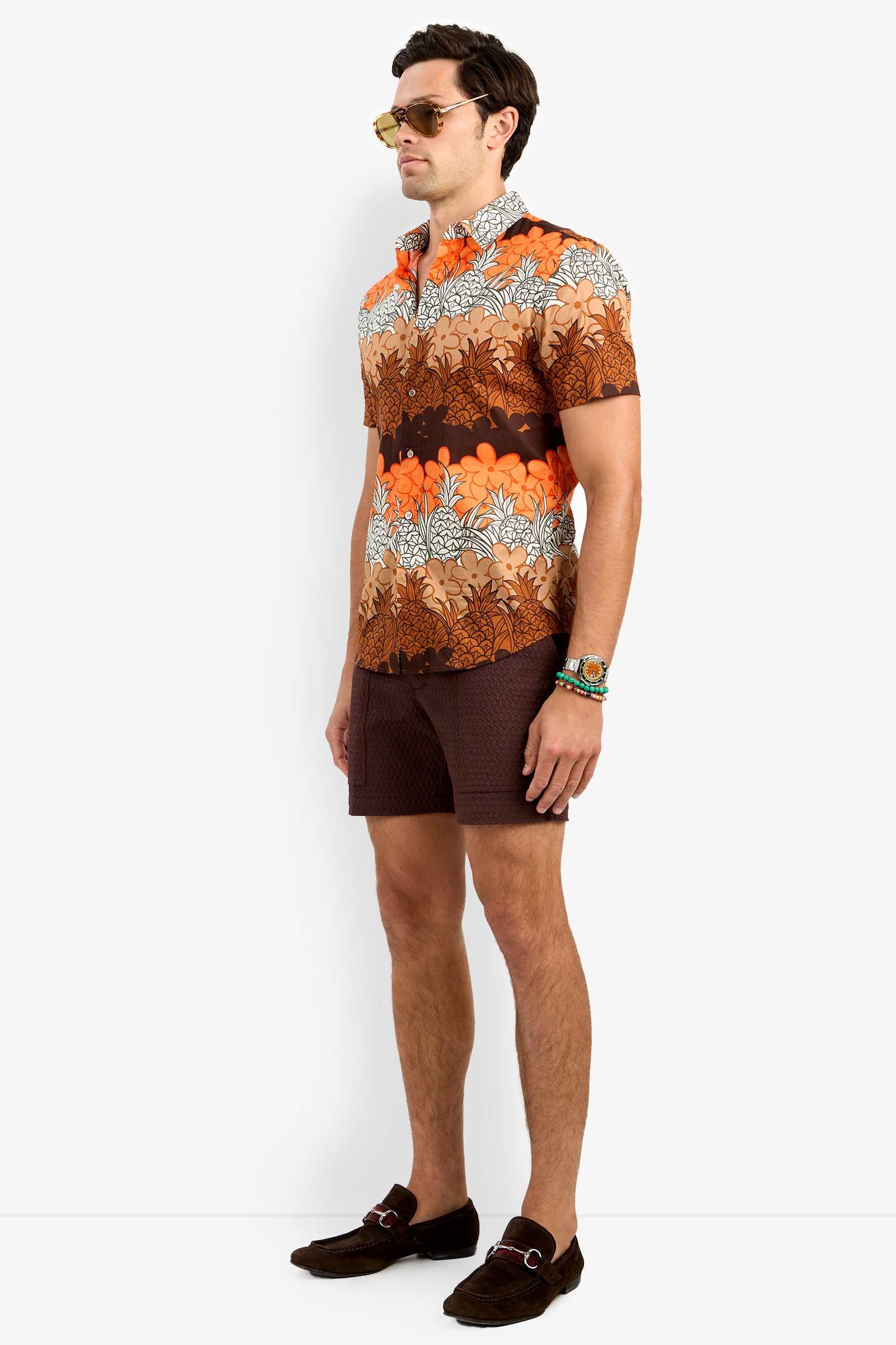 A man in sunglasses, wrist accessories, brown shorts, and loafers models the Slim Jim Shirt—a bright orange and brown floral-patterned short-sleeve—against a white background.