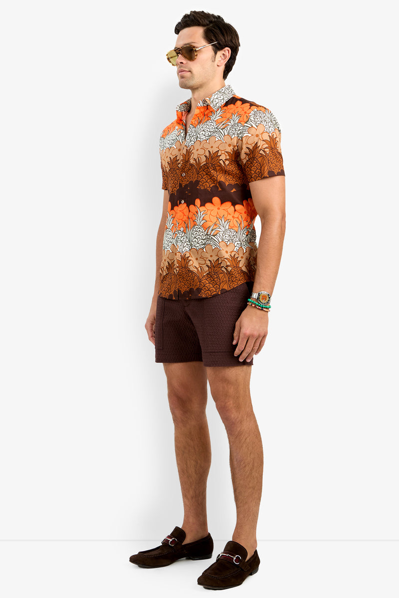 A man in sunglasses, wrist accessories, brown shorts, and loafers models the Slim Jim Shirt—a bright orange and brown floral-patterned short-sleeve—against a white background.